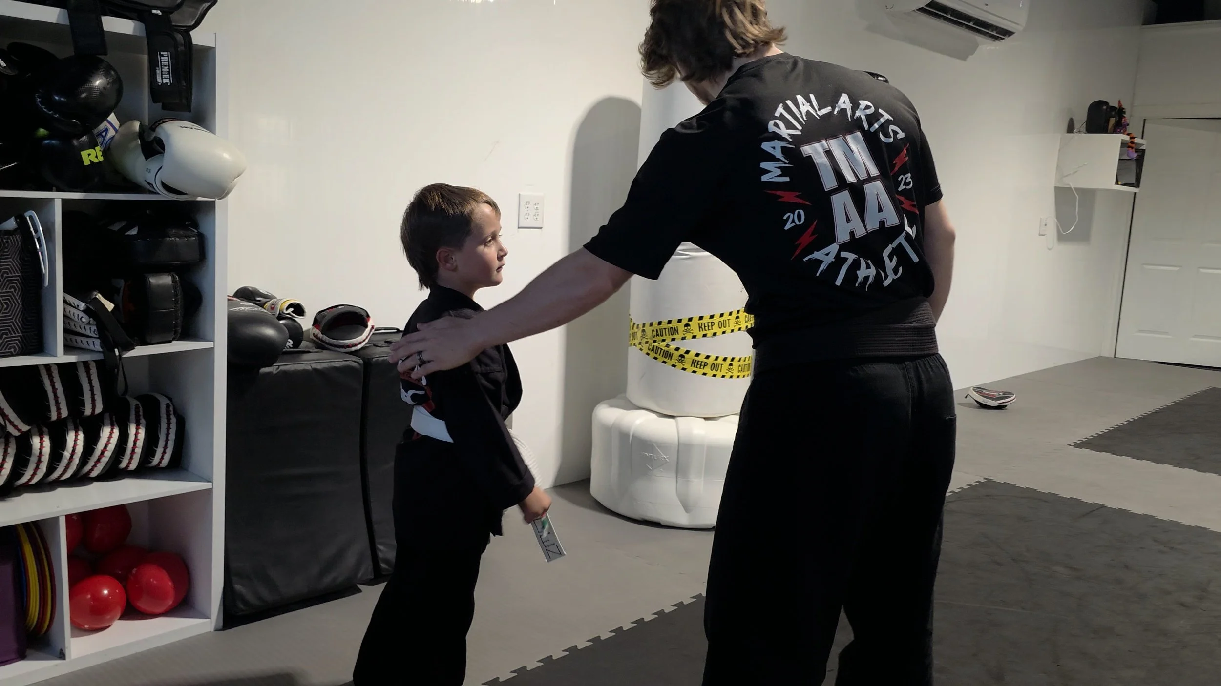 Martial arts instructor training a child in a gym with punching bags, gloves, and a taped-off practice area