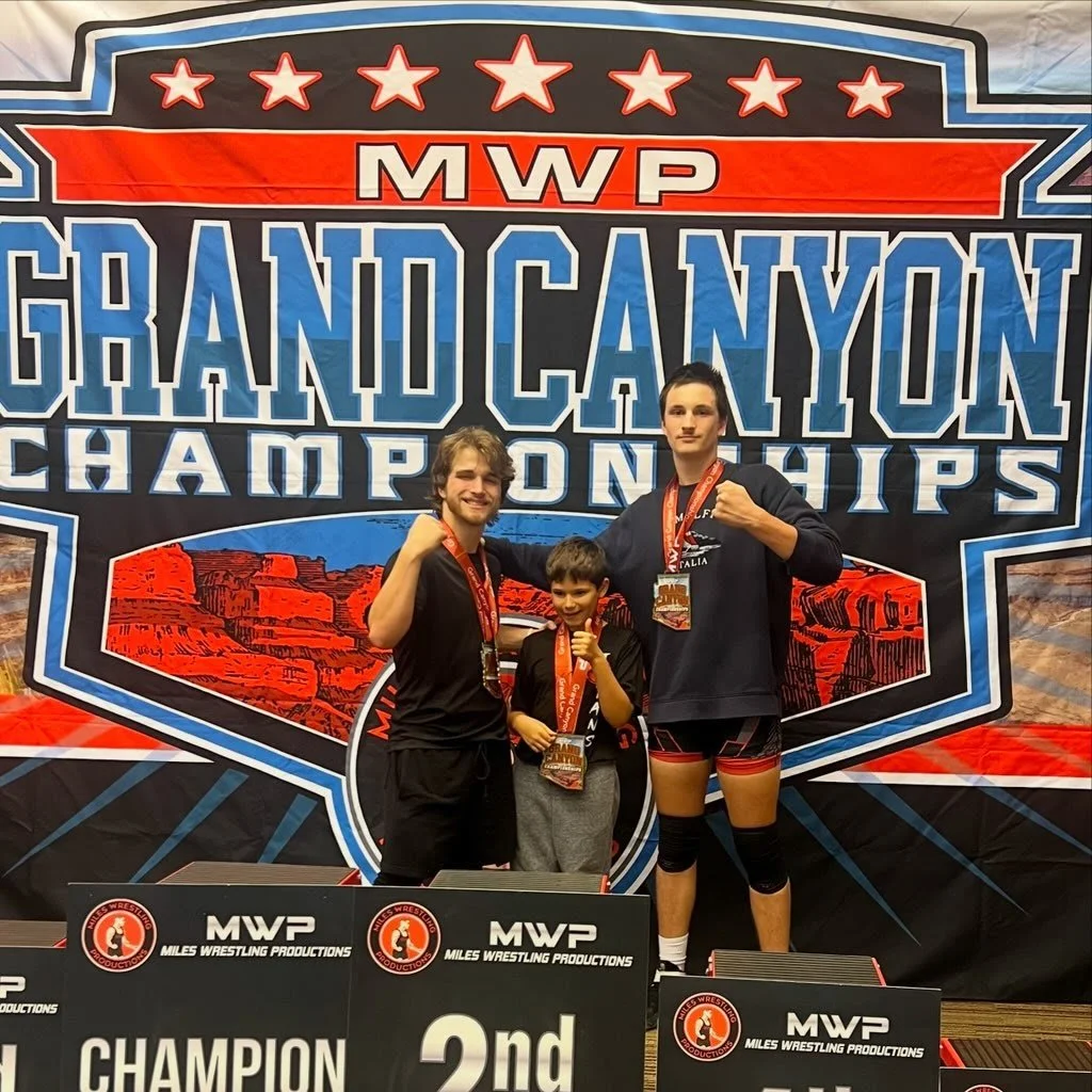 Three males posing with medals in front of a grand canyon wrestling championships banner, celebrating their wins.