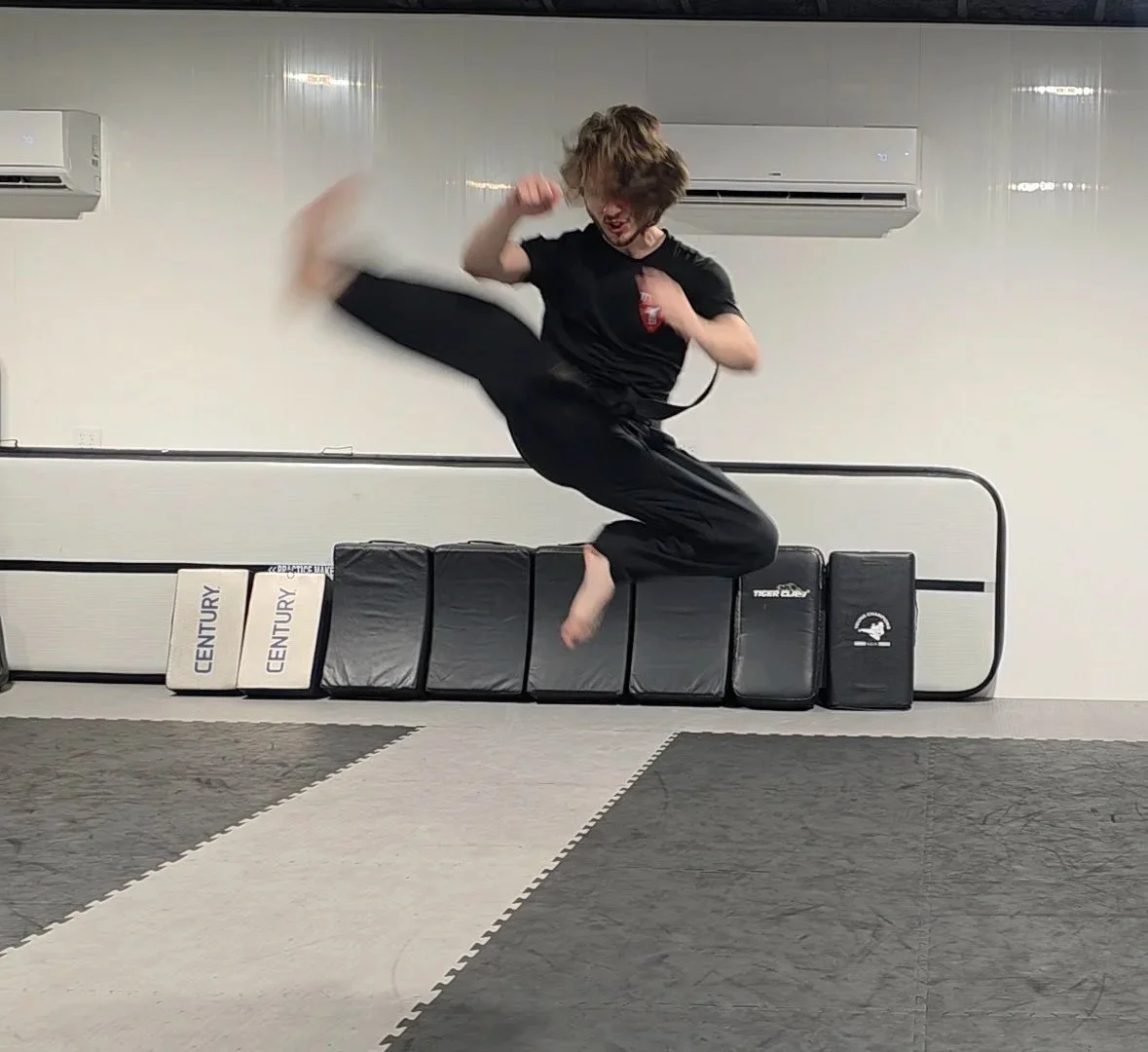 Person in mid-air performing a martial arts jump in a training room with padded mats, black and white training pads, and air conditioning units on the wall.