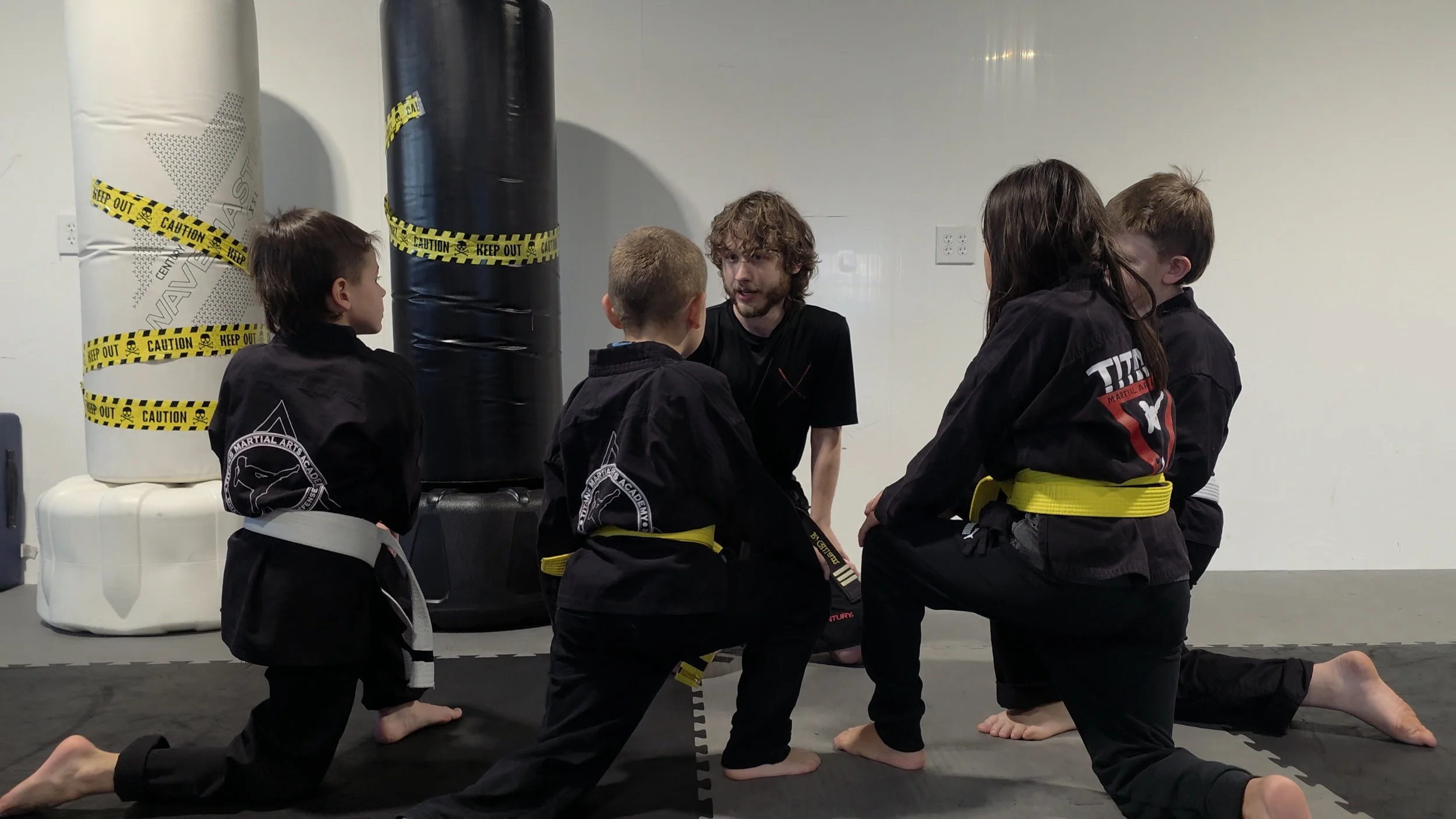 Martial arts instructor teaching children in black uniforms and colored belts on a training mat beside punching bags