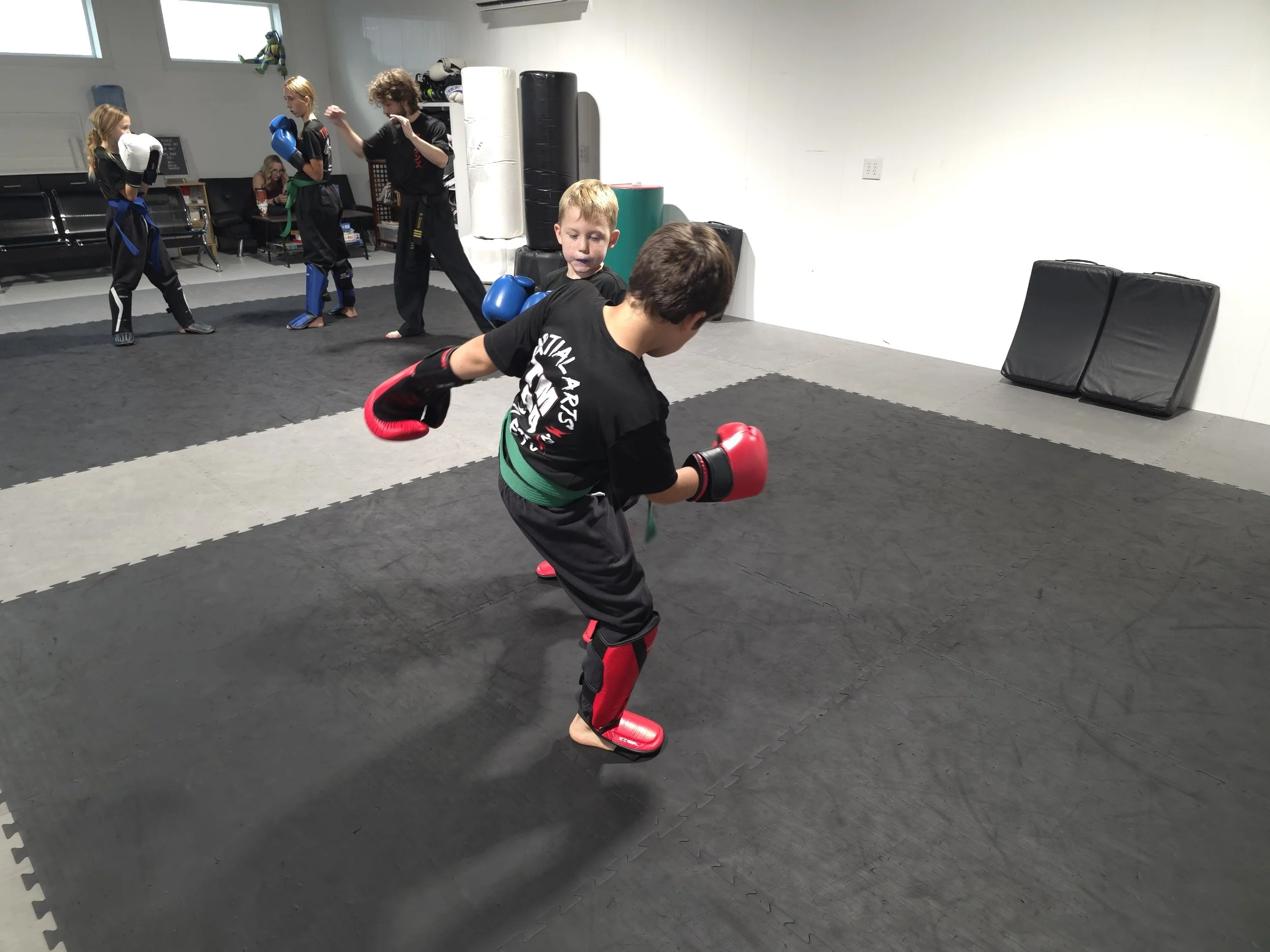 Children practicing martial arts in a dojo gym, with some wearing protective gear and sparring, on a padded floor.