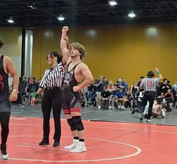 A young male wrestler with curly hair and light skin stands on a red wrestling mat with his right arm raised in victory, while a referee in a black and white striped shirt stands nearby. There are other wrestlers and spectators in the background.