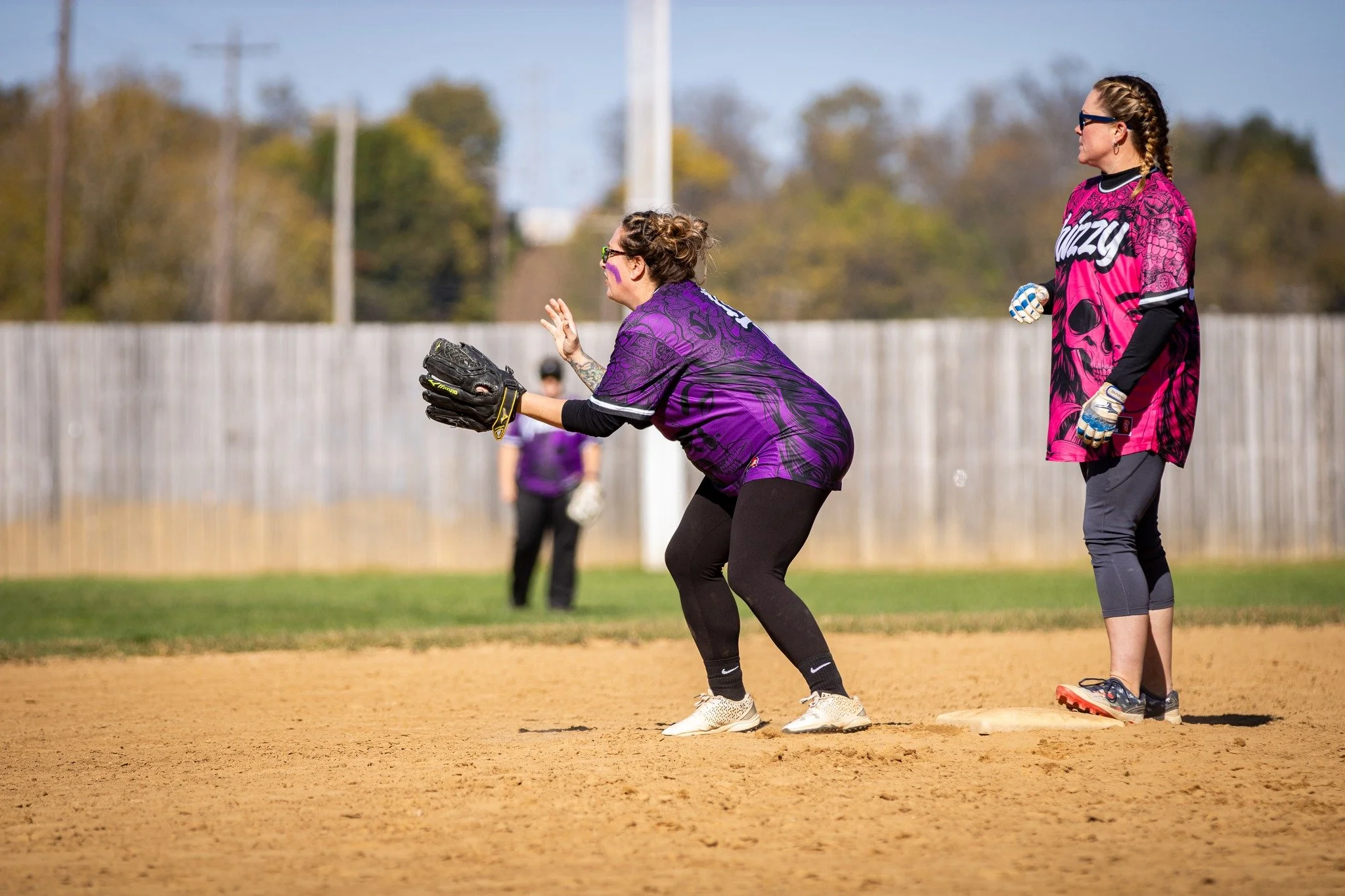 Softball players on the field wearing purple and pink jerseys during a game, focusing on one player at a base with a glove ready for action.