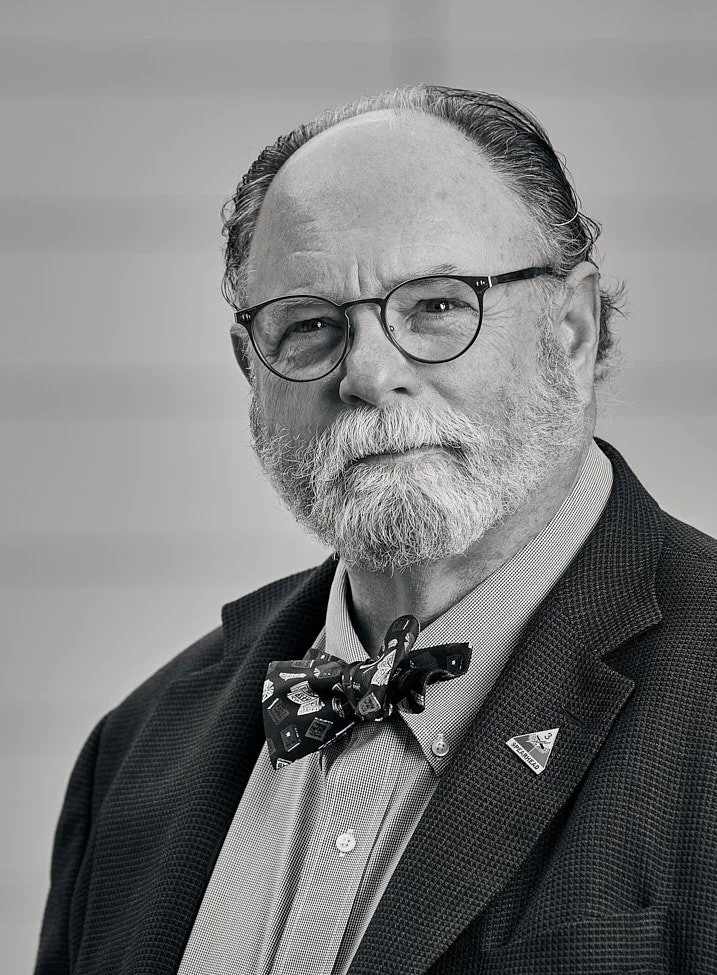 Black and white portrait of JOHN SLEMP with glasses, a beard, dressed in a suit with a bow tie, and wearing a pin on his lapel.