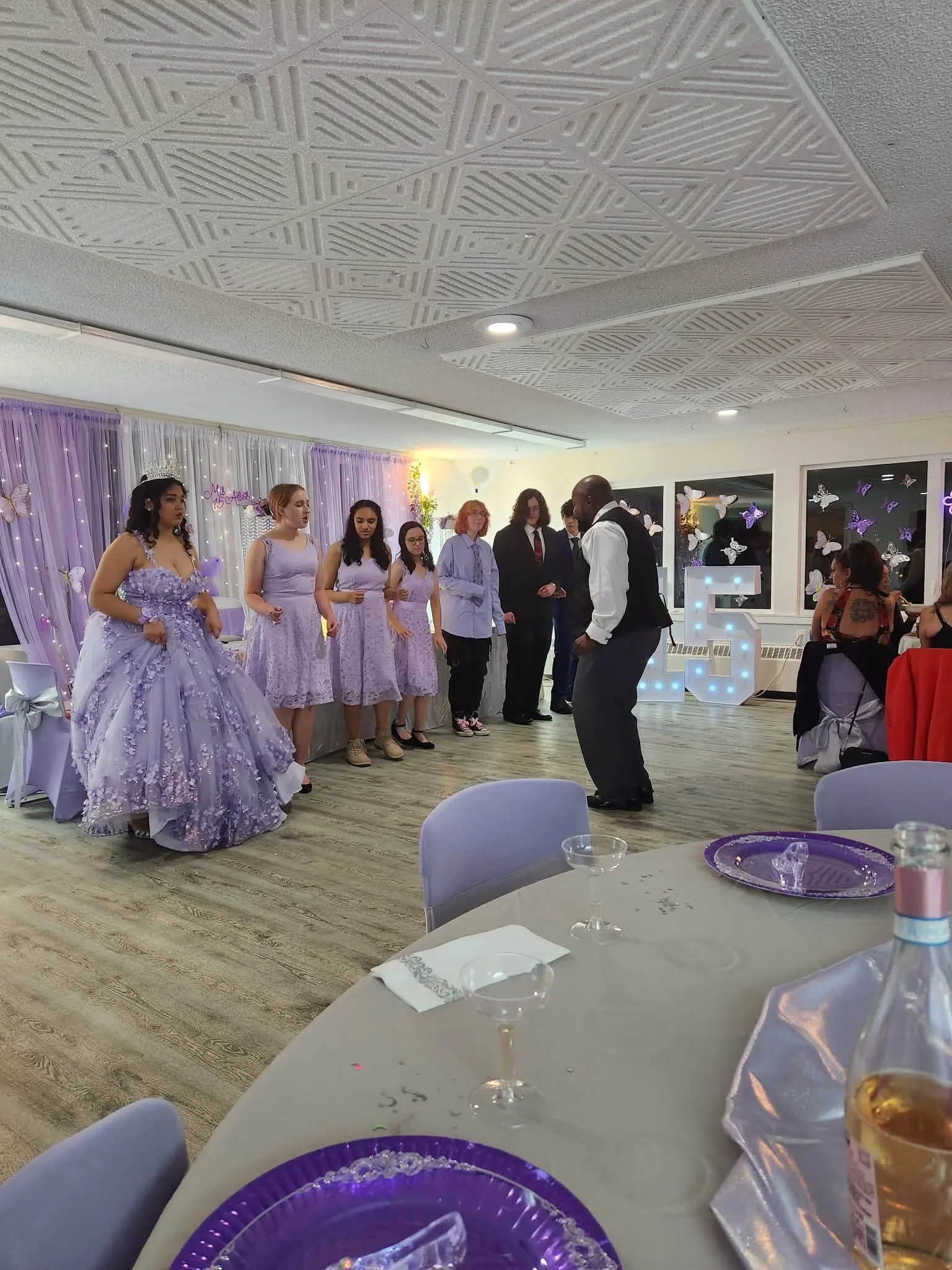 People at a celebration event, some dressed in purple gowns, with a decorated backdrop, a lit number 5, and tables with drinkware and purple plates.