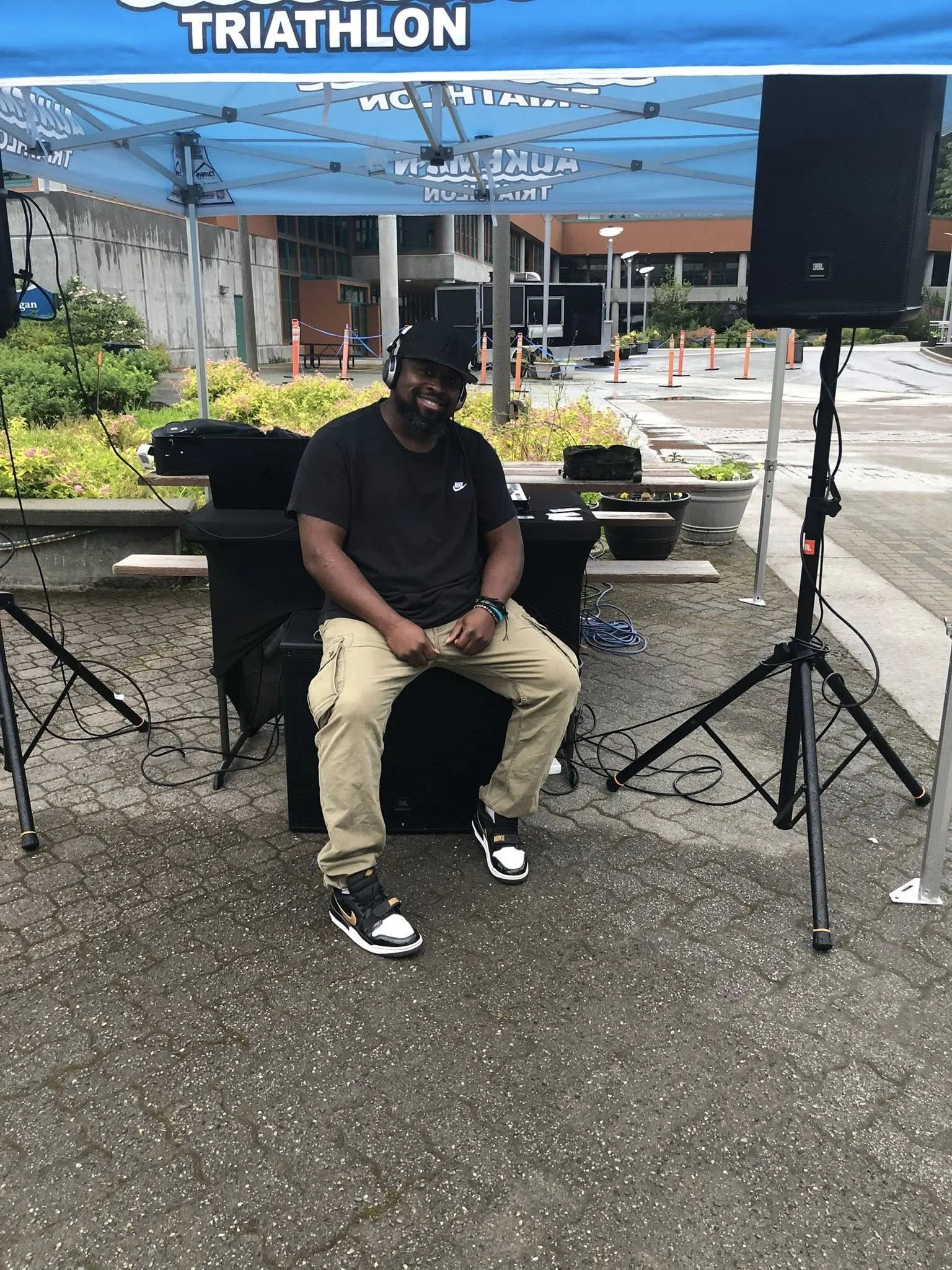 A man sitting under a blue tent at an outdoor event, wearing a black t-shirt, beige pants, and black and white sneakers, with headphones on, smiling at the camera.