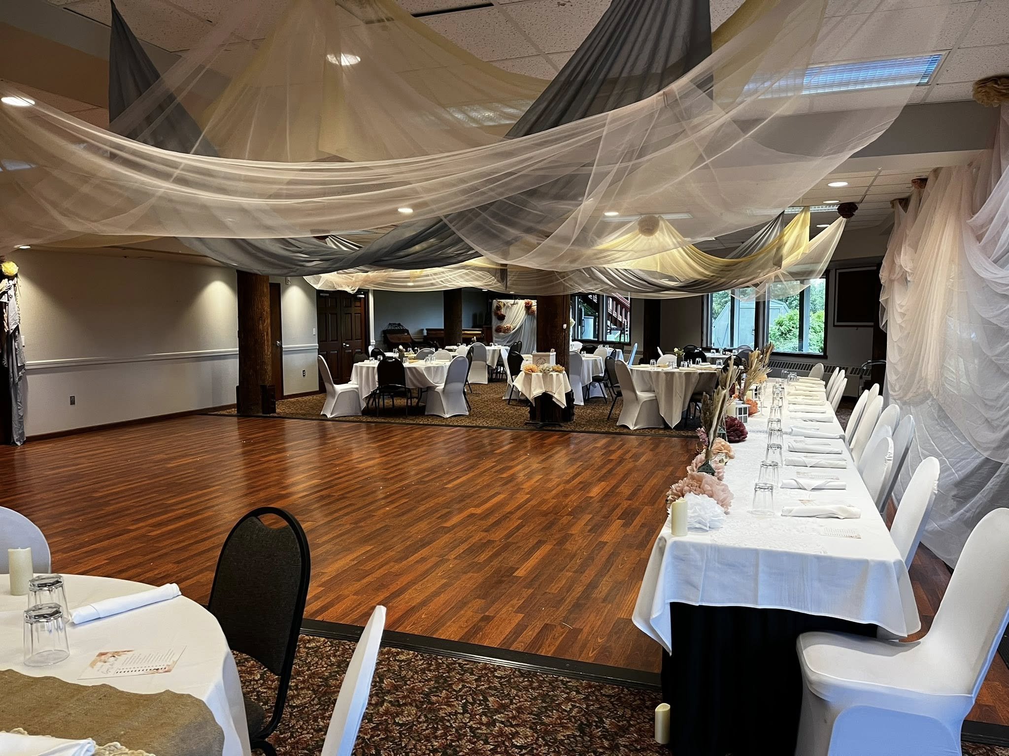 An event space decorated for a wedding reception with draped fabric on the ceiling, round and rectangular tables with white tablecloths, chairs with white covers, and a long head table decorated with flowers.