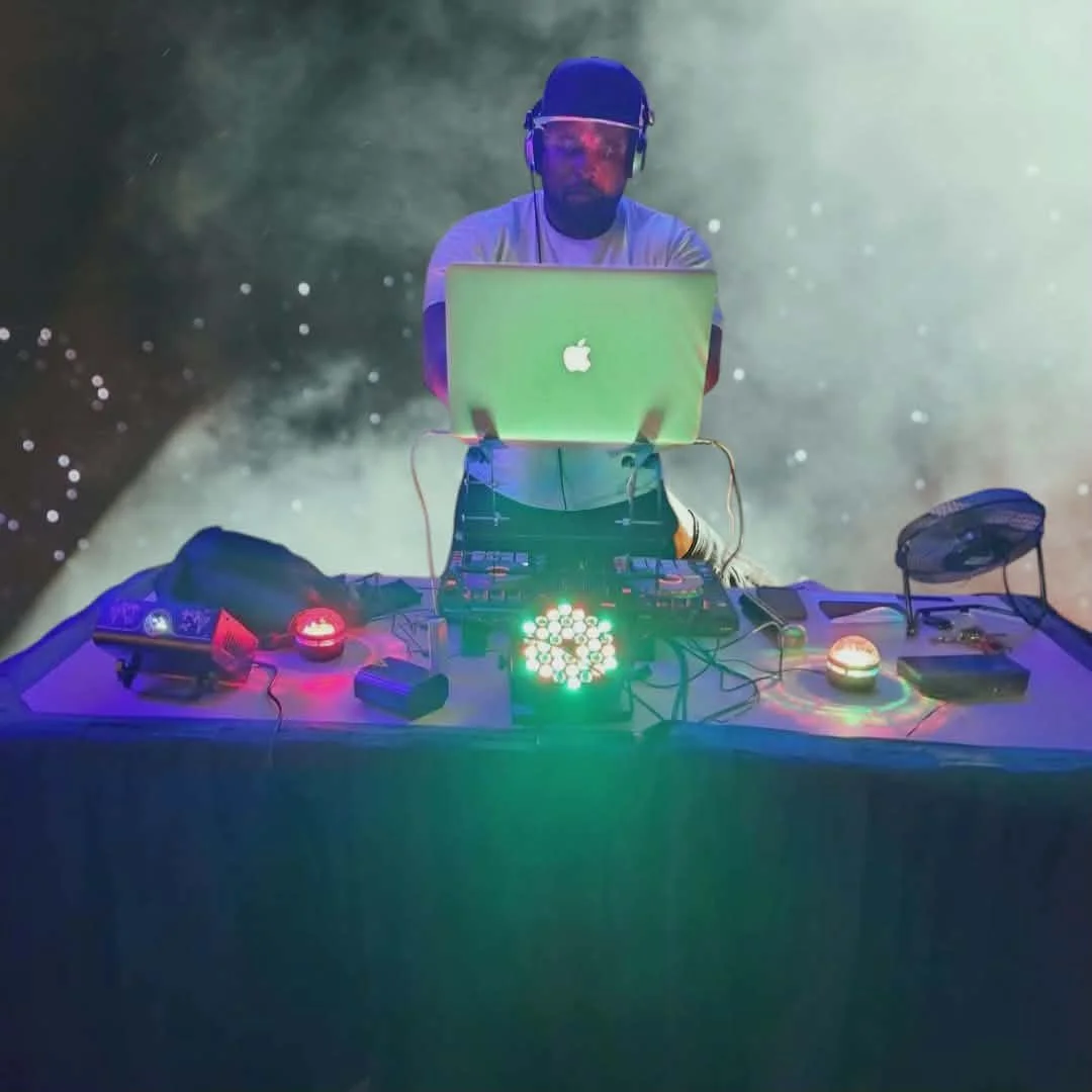 DJ Al Boogie (Al Wilson) wearing headphones and sunglasses, working on a laptop with an Apple logo, surrounded by colorful DJ equipment and lighting, in a space-themed background.
