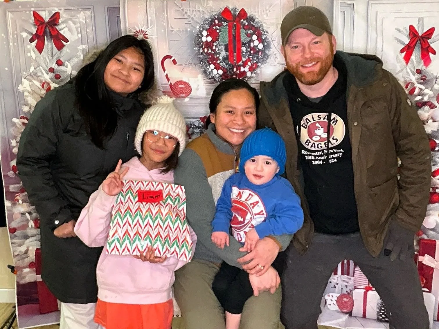 A group of five people, including two children and three adults, posing in front of a decorated Christmas backdrop with wreaths, ribbons, and wrapped gifts.