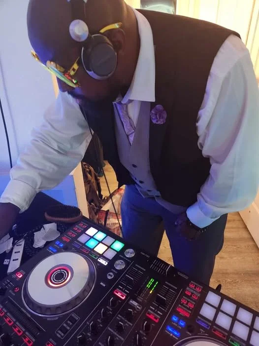 A DJ wearing headphones, a white shirt, and a black vest, standing by a DJ controller with colorful lights.