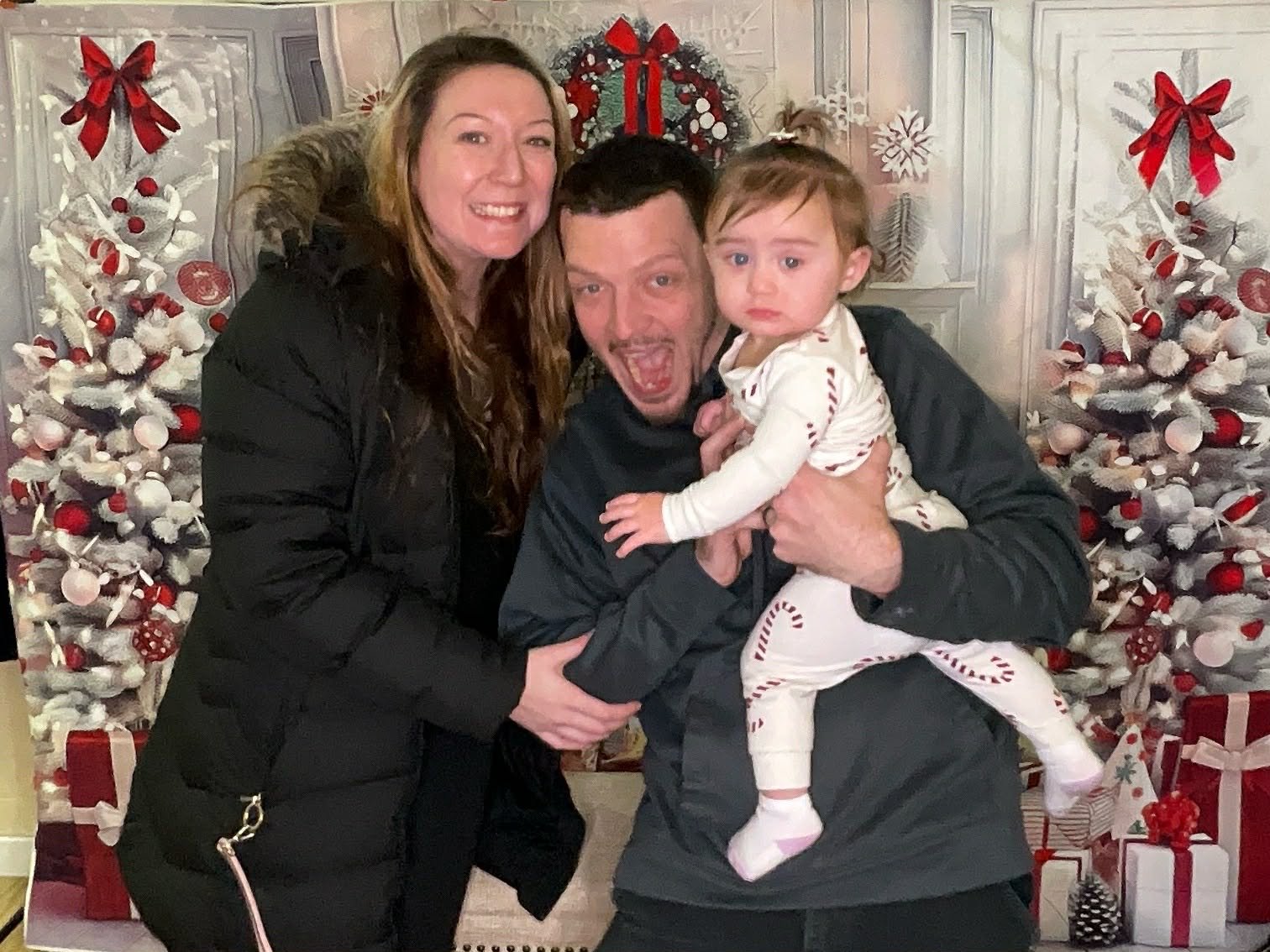 A family of three posing in front of decorated Christmas trees with presents, with a woman on the left, a man in the middle holding a baby girl on the right.