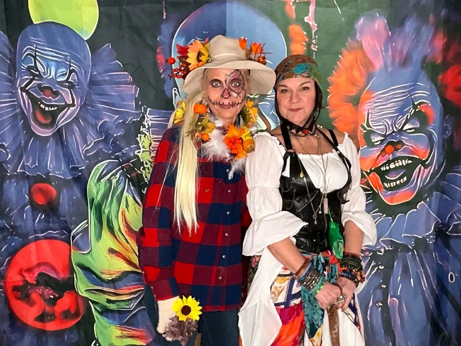 Two women in festive costumes standing in front of a vibrant clown-themed backdrop. One woman is dressed as a colorful scarecrow with face paint, a large hat, and sunflowers. The other woman wears a pirate or gypsy-inspired outfit with layered jewelr