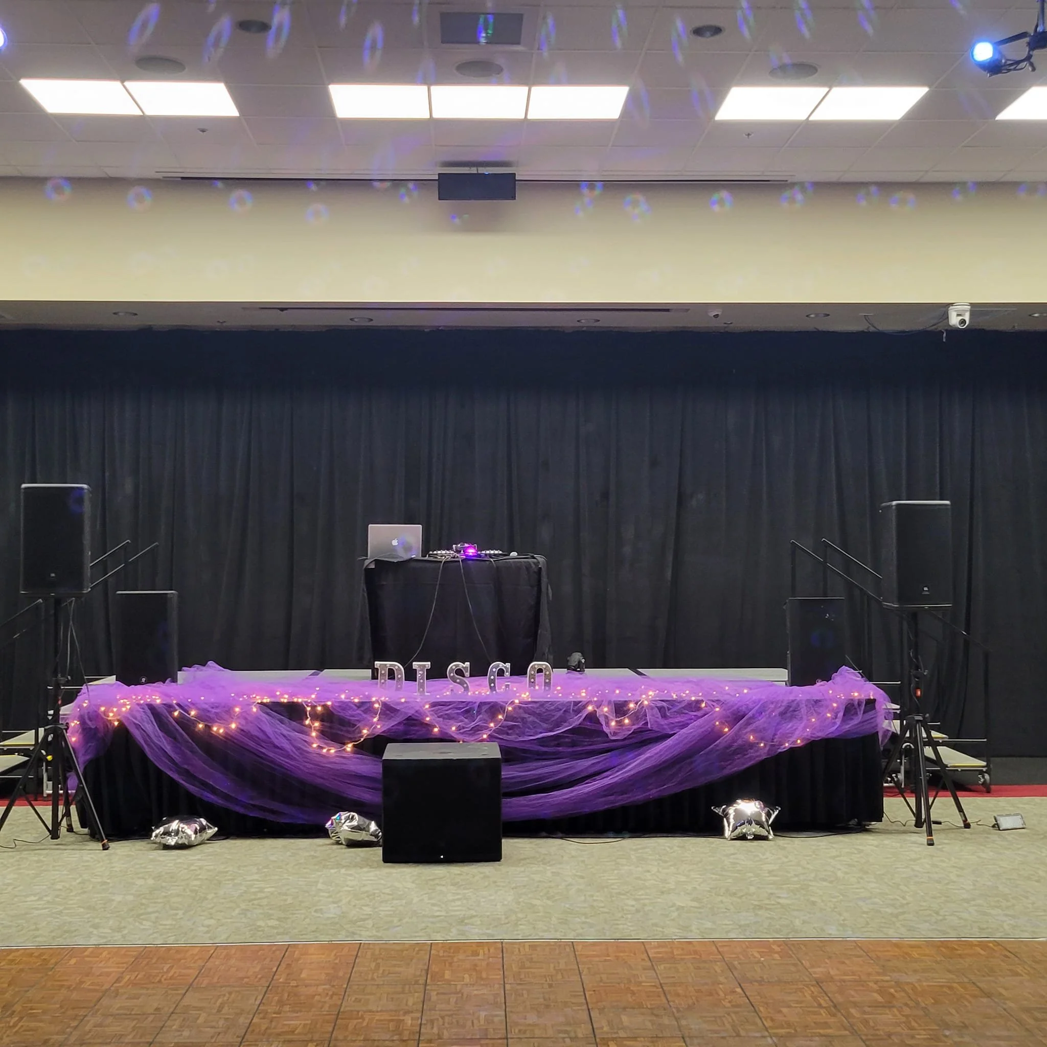 Empty party stage decorated with purple tulle, fairy lights, and star-shaped balloons, with DJ equipment and speakers set up, against a black curtain backdrop.