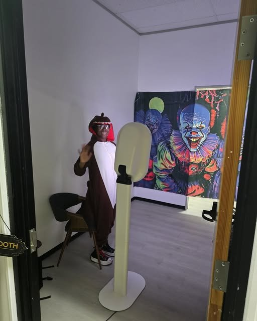 A person dressed in a teddy bear costume standing in a photo booth, waving and smiling. Behind them is a colorful clown-themed painting.