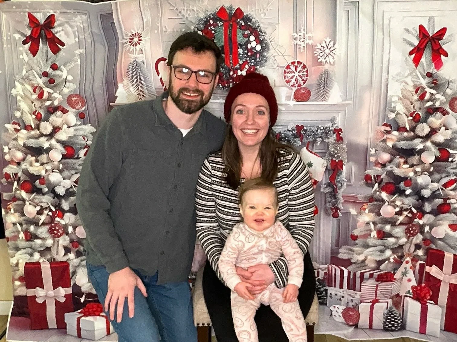 A family of three posing in front of a festive Christmas backdrop with decorated white Christmas trees, a decorated fireplace, and wrapped gifts. The family includes a man with glasses, a woman with long hair wearing a red hat, and a smiling baby sit