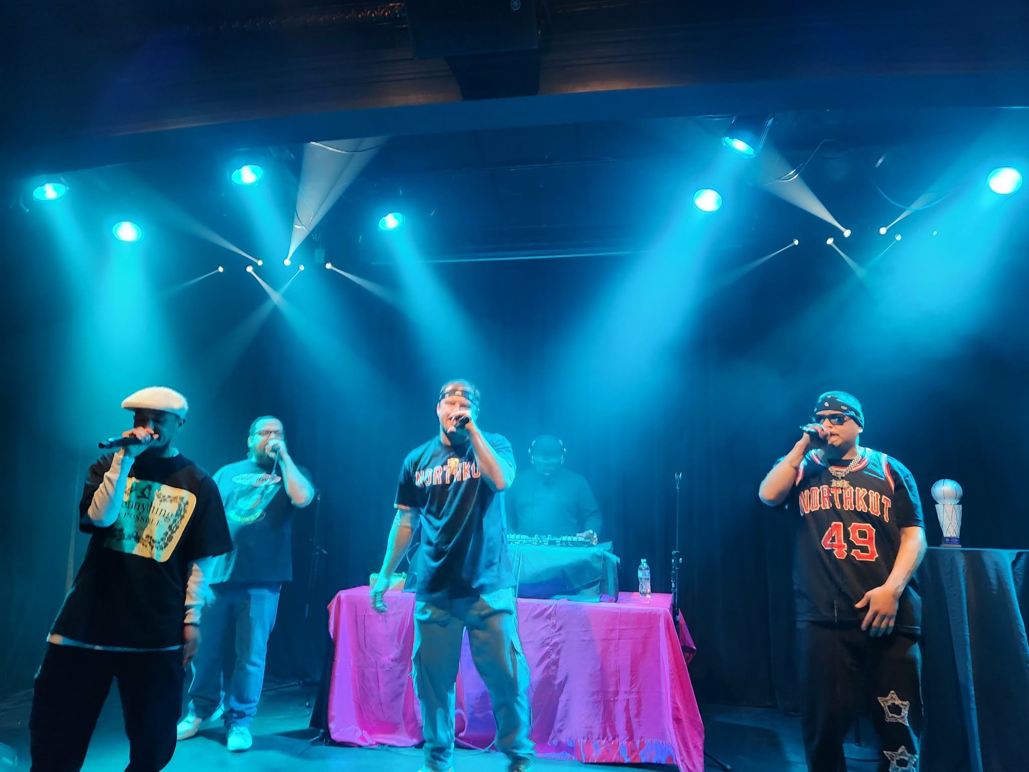 Four men performing on stage with blue stage lighting, one with a microphone, one with a DJ setup, and two wearing casual streetwear, including branded shirts and accessories.