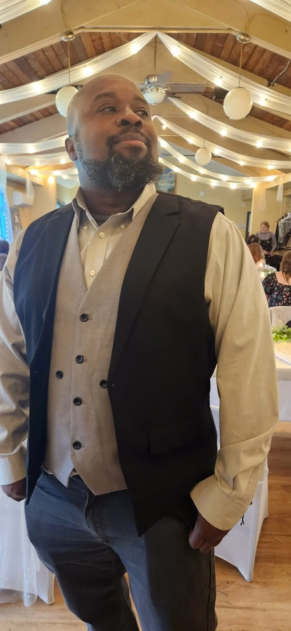 DJ Al Boogie (Al Wilson) A man with a beard dressed in a beige vest and shirt, with a black vest over it, at a decorated indoor event with string lights and round white lanterns hanging from the ceiling, and people seated at tables in the background.