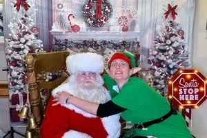 A woman dressed as an elf hugging Santa Claus in a festive holiday setting with decorated trees and a 'Santa Stop Here' sign.