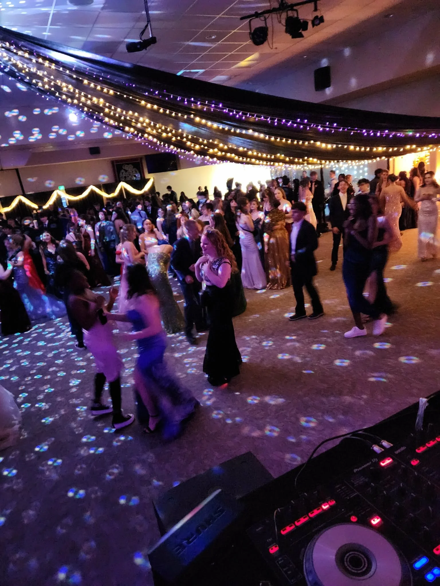 Crowd of young people dancing at a dance party with colorful lights and string lights decorating the ceiling.