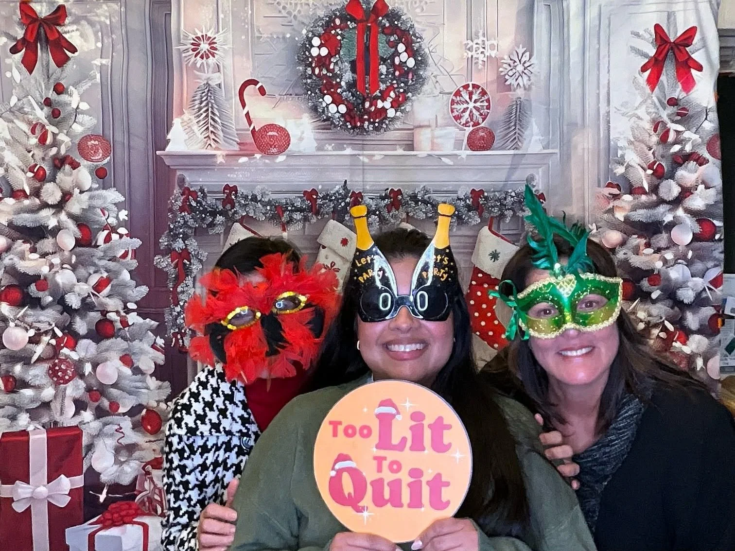 Three women wearing colorful masks posing in front of a decorated fireplace and Christmas trees with presents on Christmas.