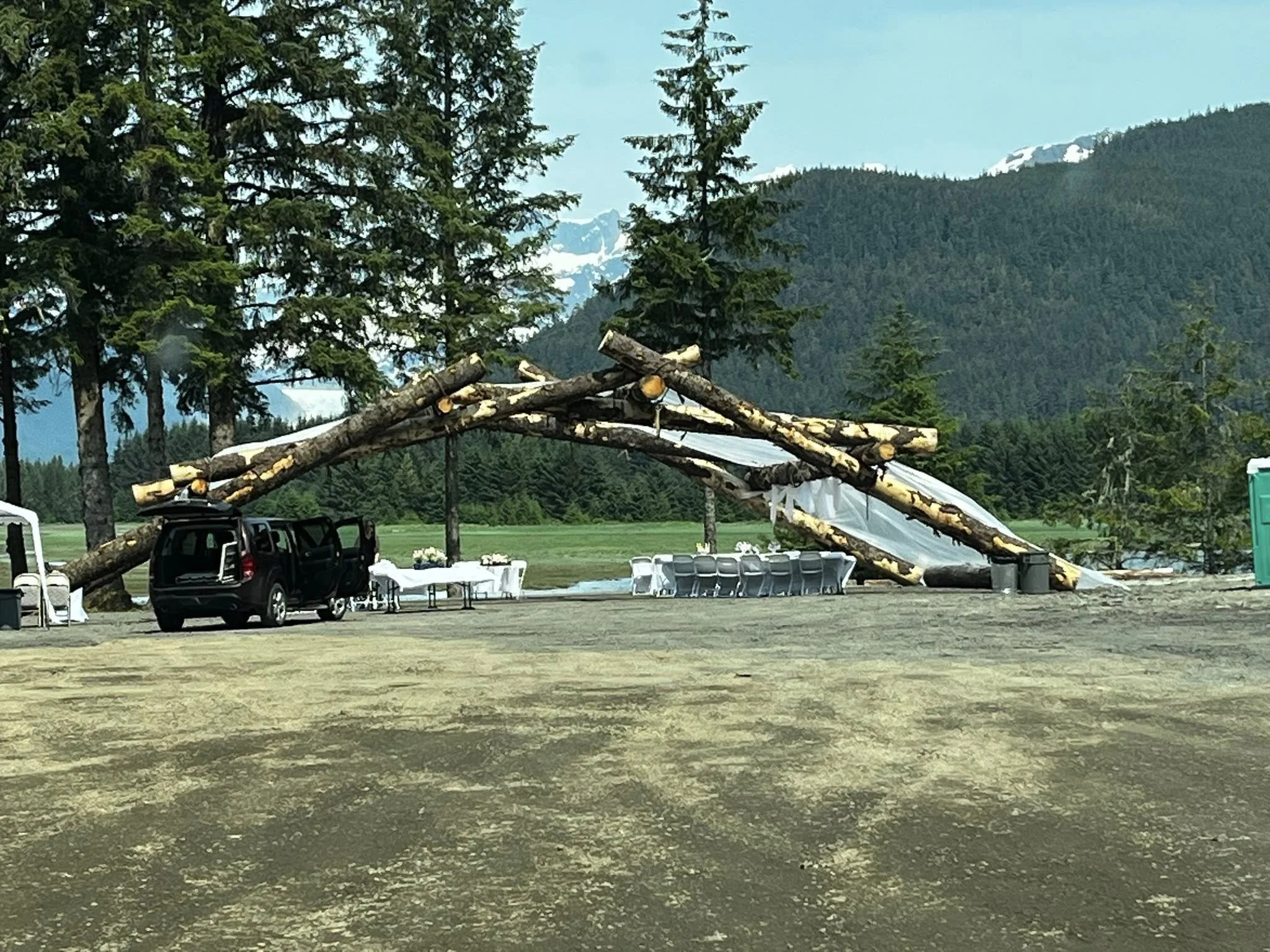 Outdoor event setup with large wooden log arch and tables in a natural mountainous setting.