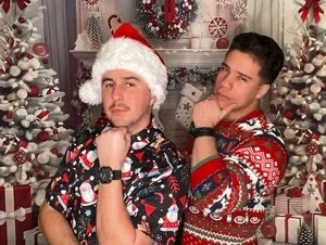Two young men posing in front of Christmas decorations, one wearing a Santa hat and a holiday-themed shirt, the other in a colorful Christmas sweater, both with thoughtful expressions.