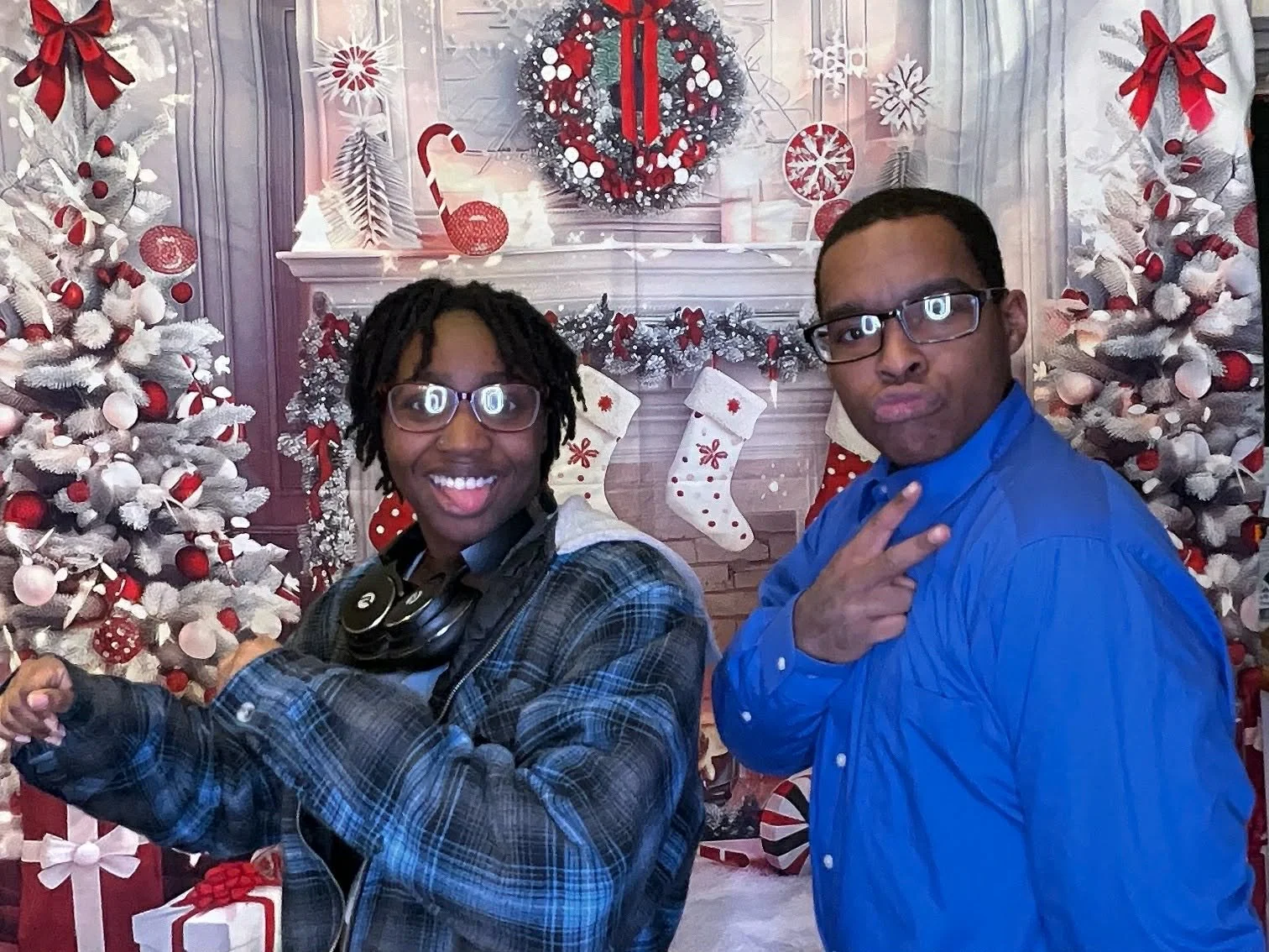 Two individuals posing in front of a Christmas-themed backdrop with decorated white trees, stockings, and a wreath, celebrating the holiday season.