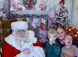 A person dressed as Santa Claus sitting with three young children in a holiday-themed room with Christmas decorations, including a white Christmas tree and stockings.