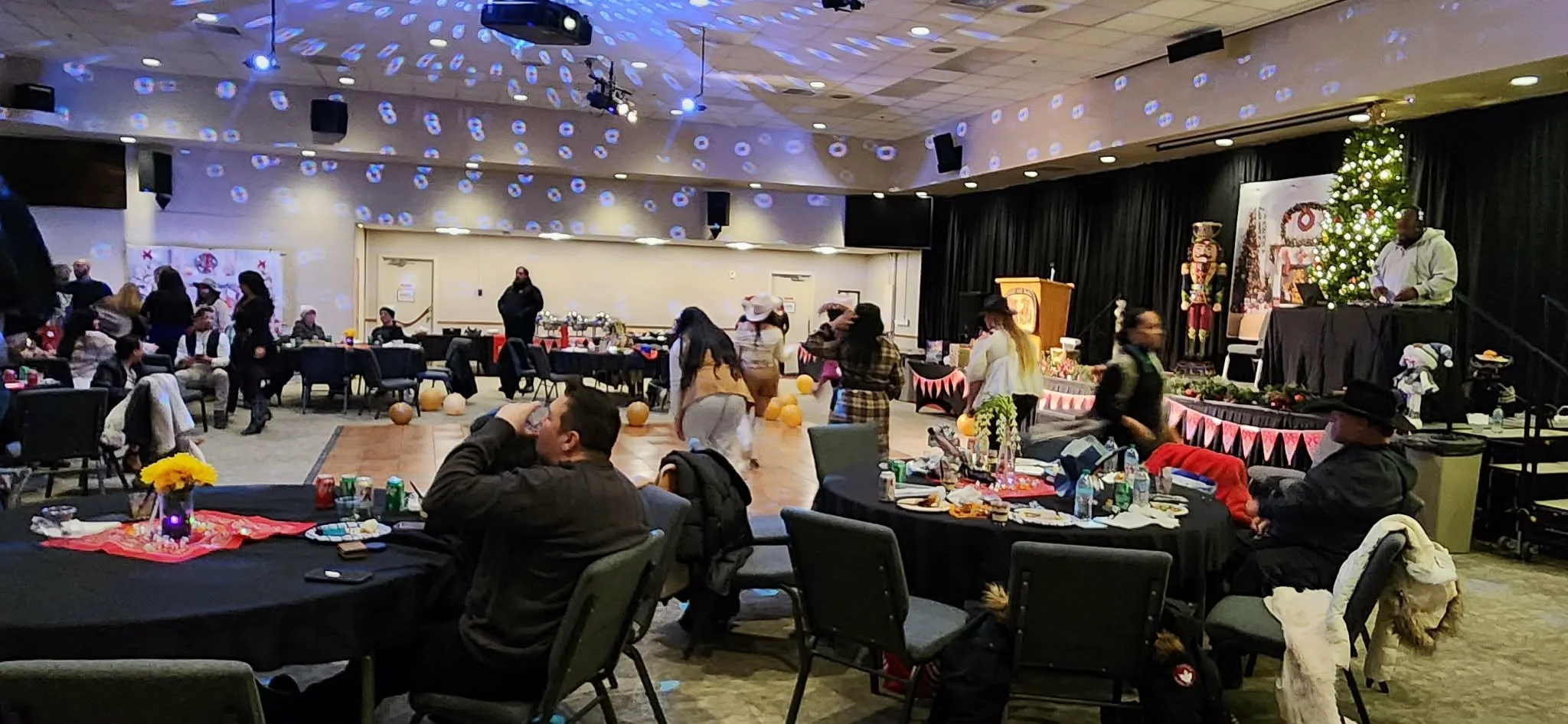 Indoor holiday party with people dancing on a dance floor, decorated with Halloween and Christmas decorations, tables with food and drinks, a DJ or musician on stage, and festive lights and ornaments.
