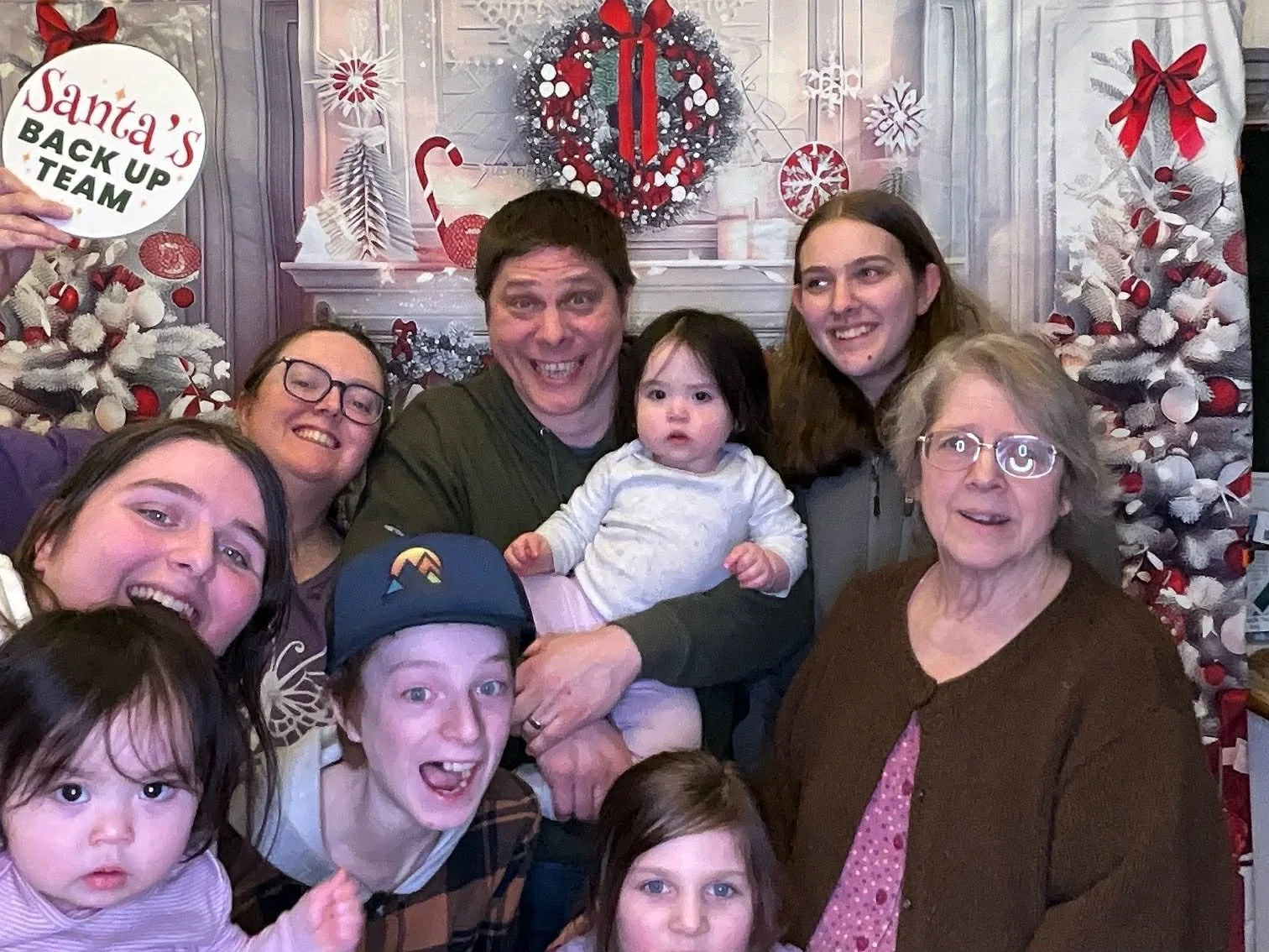 Family group photo celebrating Christmas in front of festive holiday decorations, including a fireplace decorated with red and white wreaths, ornaments, and bows. One person is holding a sign that says 'Santa's Backup Team'.