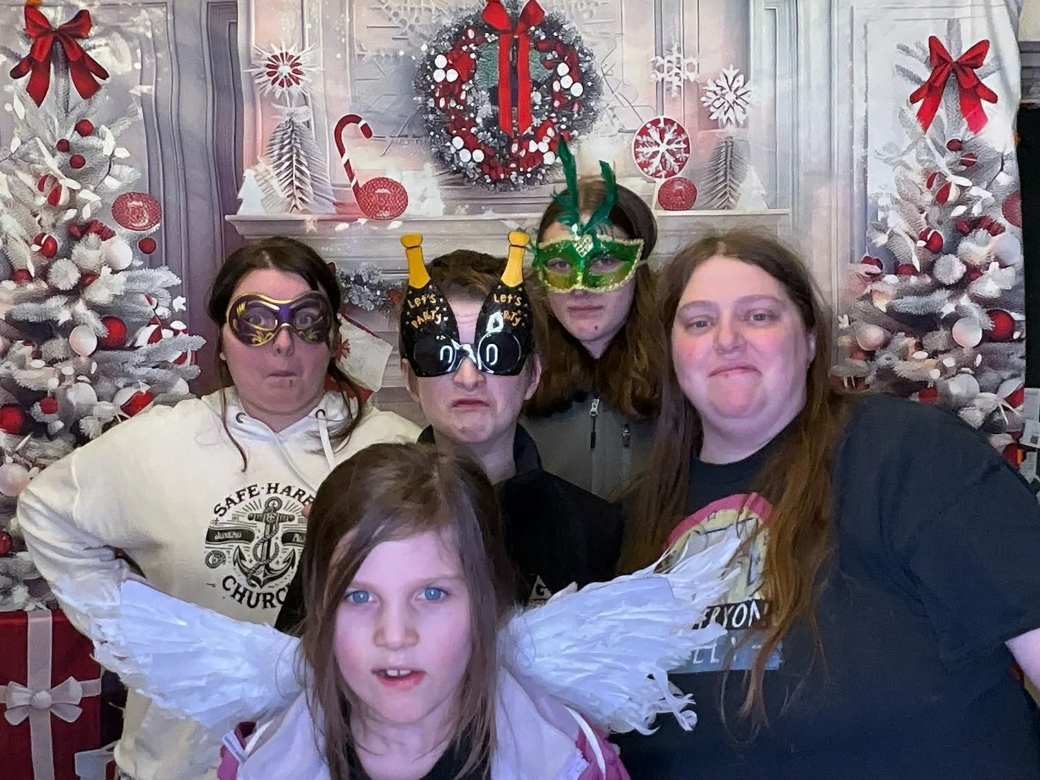 Group of five people with humorous glasses and masks posing in front of Christmas decorations, including a white Christmas tree with red and white ornaments, a red and white wreath, and wrapped presents.