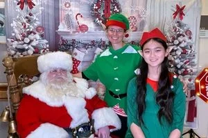 A person dressed as Santa Claus sitting next to two women dressed as elves, with Christmas decorations and a decorated tree in the background.