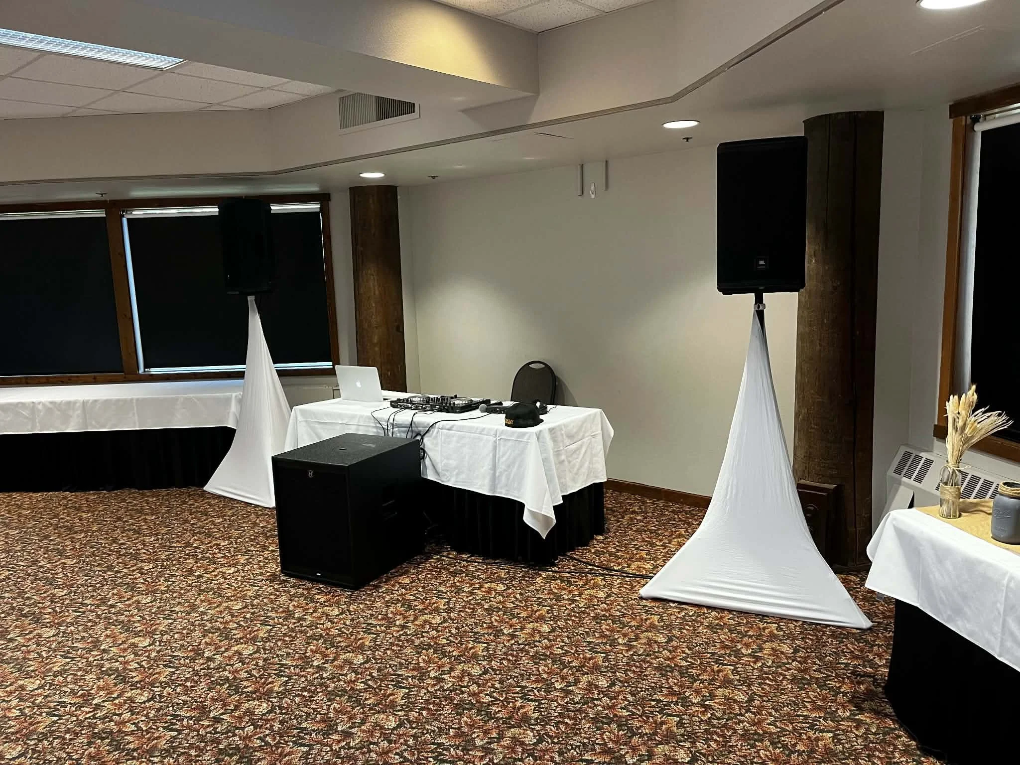 An indoor event space with DJ equipment including speakers, a laptop, and a mixer on a table covered with a white tablecloth. There are black speakers on stands with white covers, and black cables on the table. The room has patterned carpet, windows 