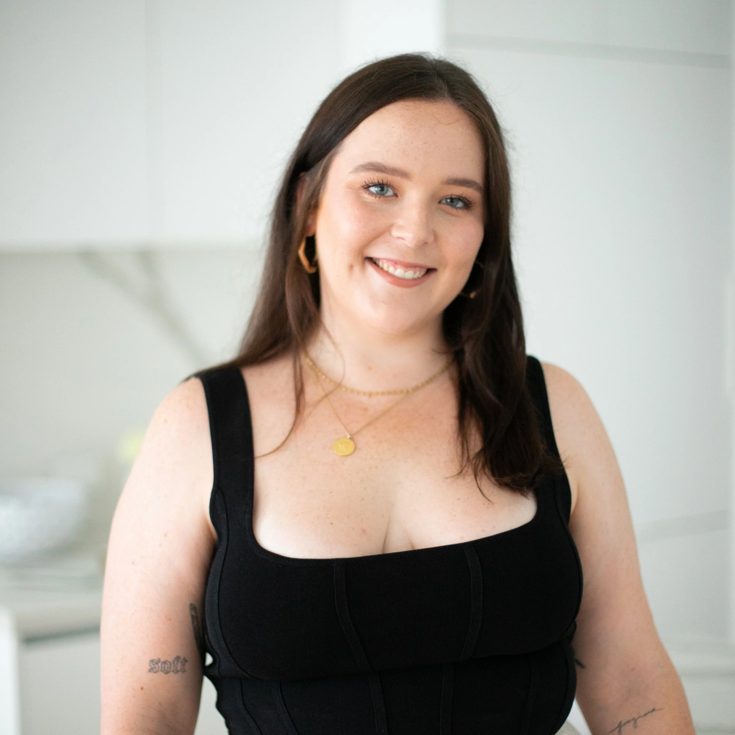 Smiling woman in black dress with gold necklace and earrings standing indoors.
