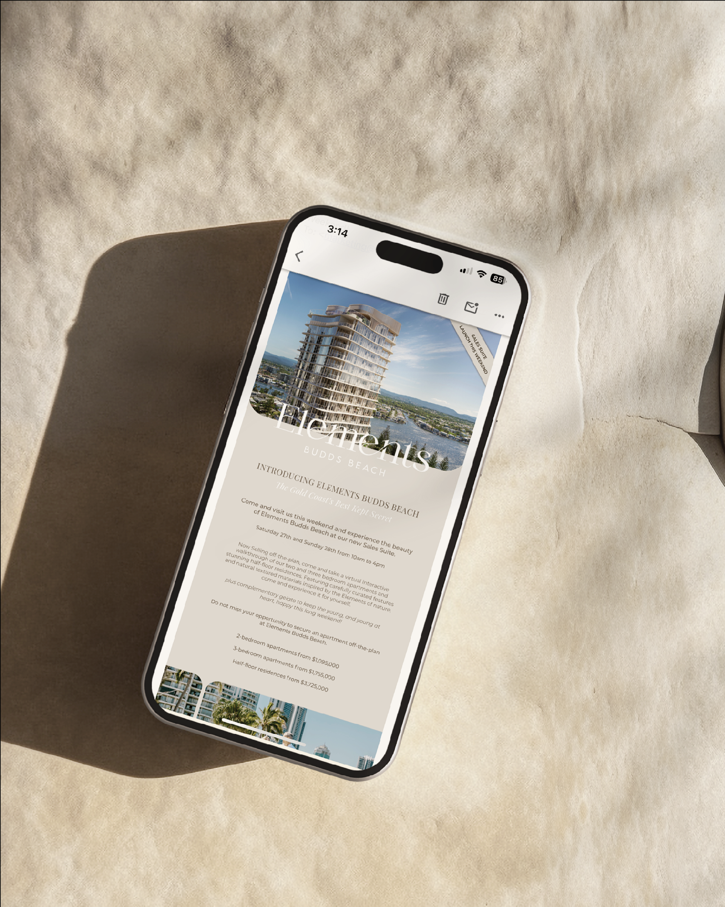 A smartphone displaying a real estate advertisement for 'Elements Budds Beach' showing a high-rise building and text details. The phone is placed on a textured surface, casting a shadow.