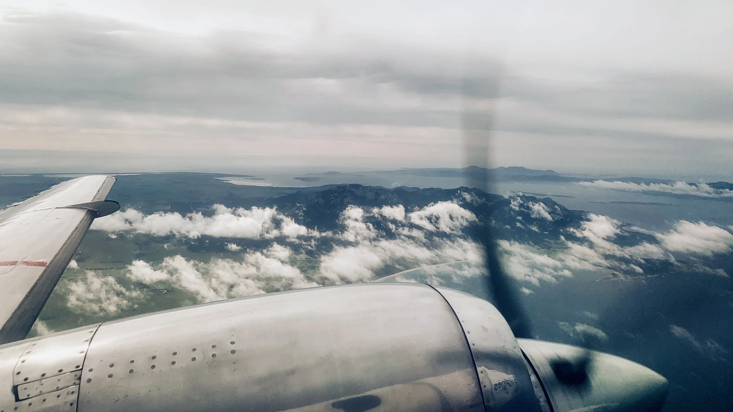 View from an airplane window showing the airplane's engine and wing, with clouds, mountainous terrain, and a body of water below under a cloudy sky.