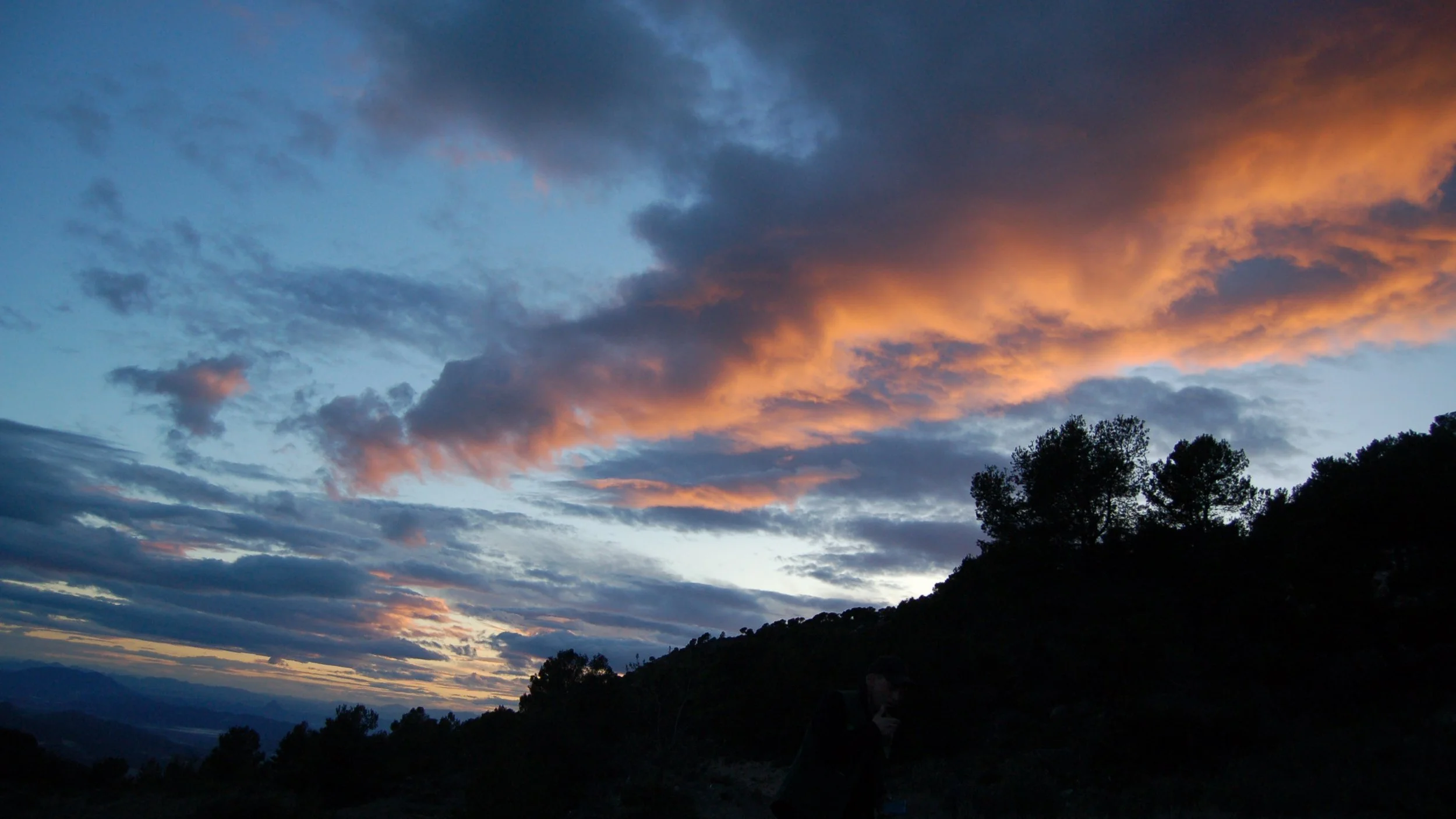 Sunset over a mountain range with dark clouds and orange-pink illuminated clouds in the sky, and silhouetted trees on a hillside.