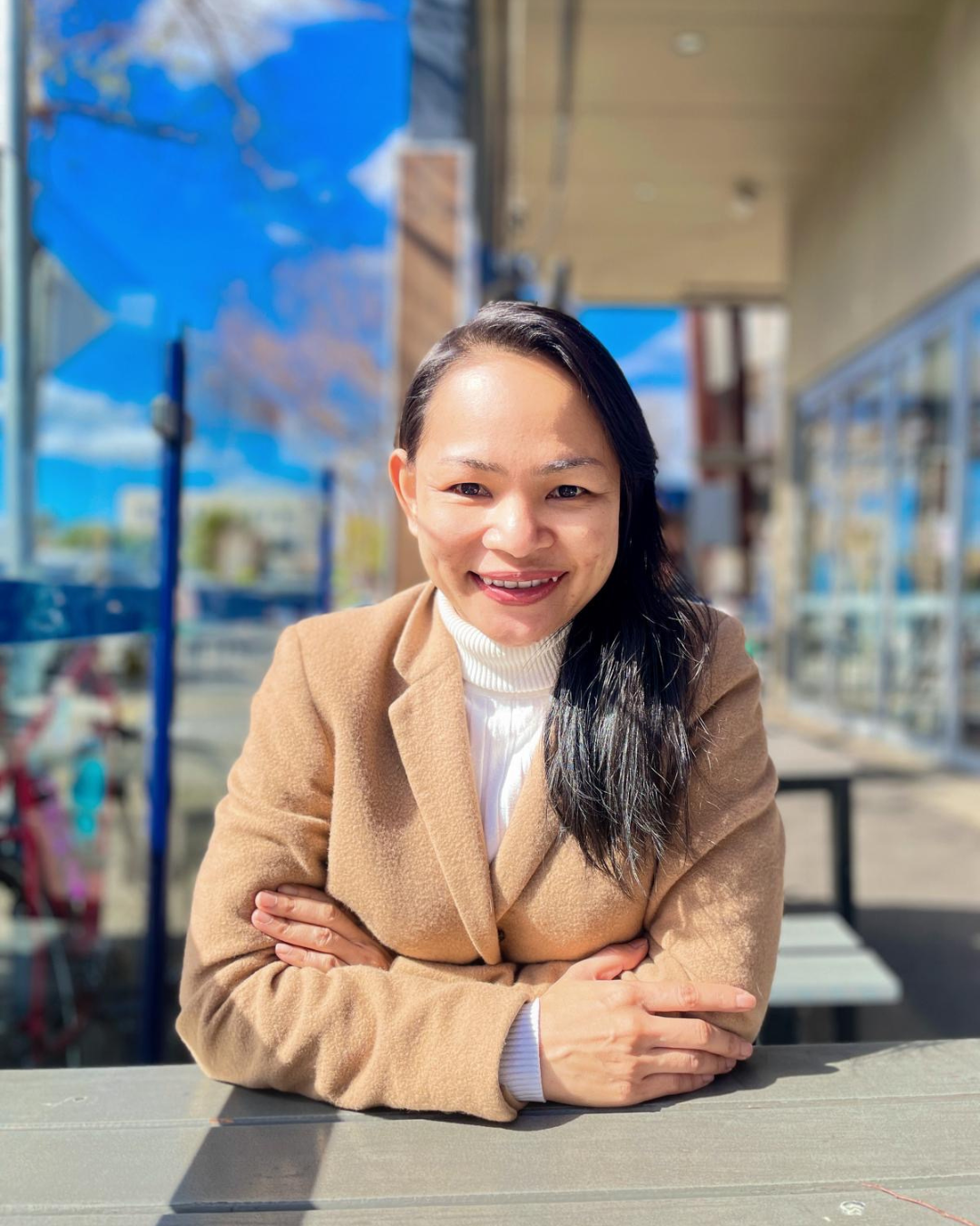 A woman sitting at an outdoor table, smiling warmly, wearing a tan coat and white top on a sunny day.