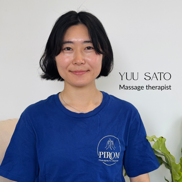 Massage therapist smiling and wearing a blue Pirom Therapeutic Massage uniform, sitting indoors with a calm and welcoming expression.