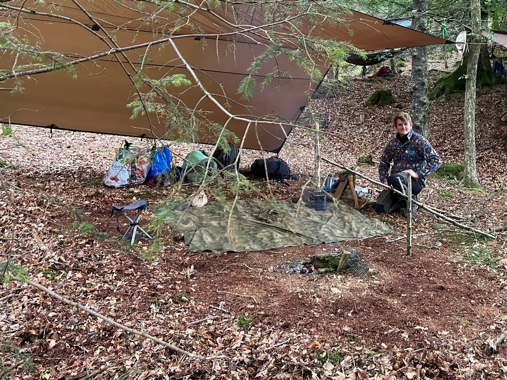 Bushcraft in the Lake District. A young man with short blonde hair sitting on the ground in a wooded area of a campsite, surrounded by backpacks, a foldable chair, and camping gear under a tarp.