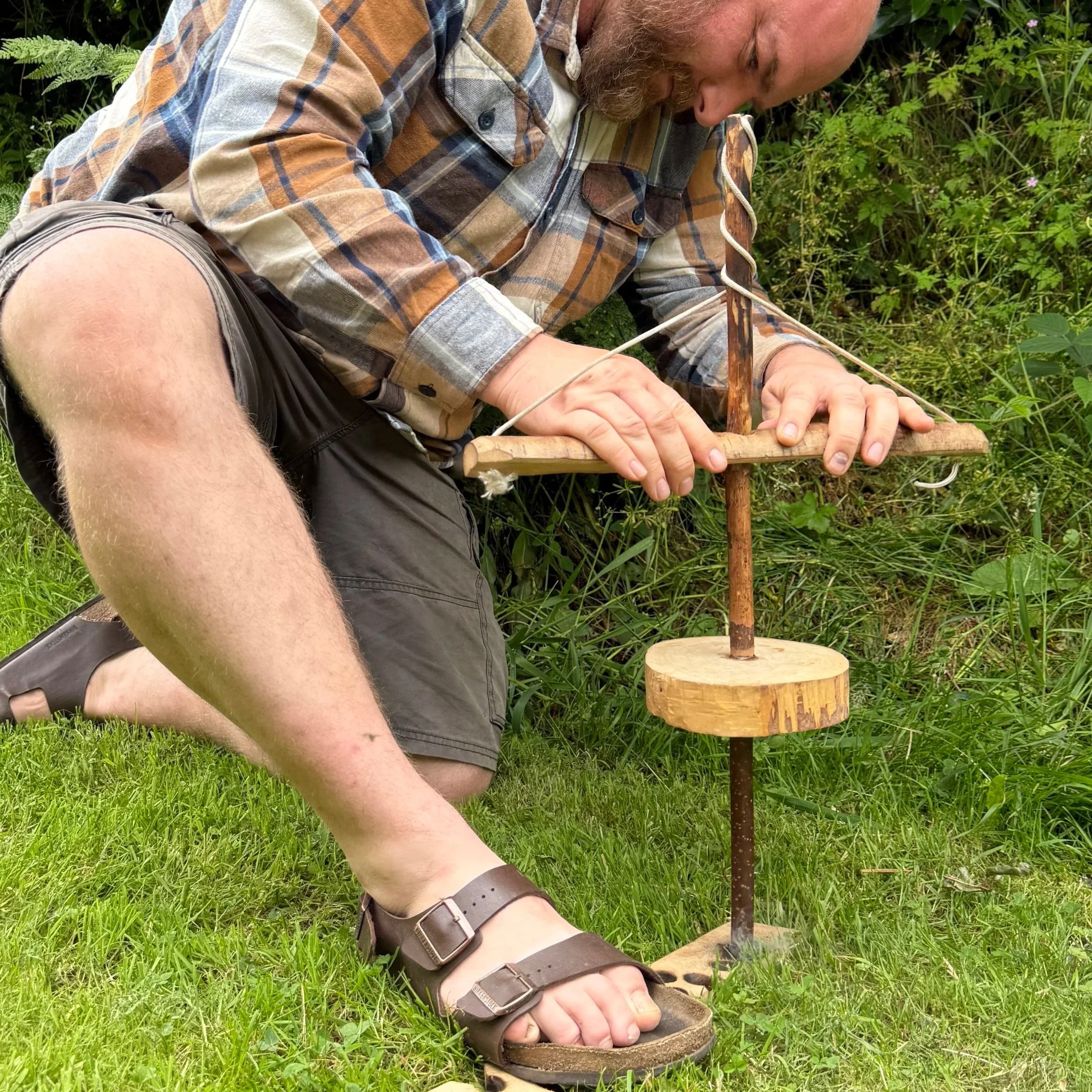 A man wearing a plaid shirt and sandals is sitting outdoors on grass, working on a small wooden project using a vertical drill press on a wooden surface, surrounded by green plants.