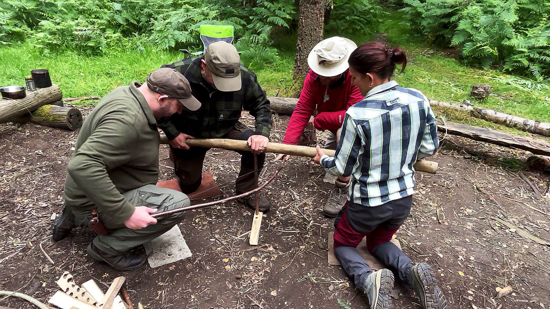 Bushcraft in the Lake District. Four people outdoors in a wooded area working together to create an ember using group bow drill friction fire, on the forest floor.