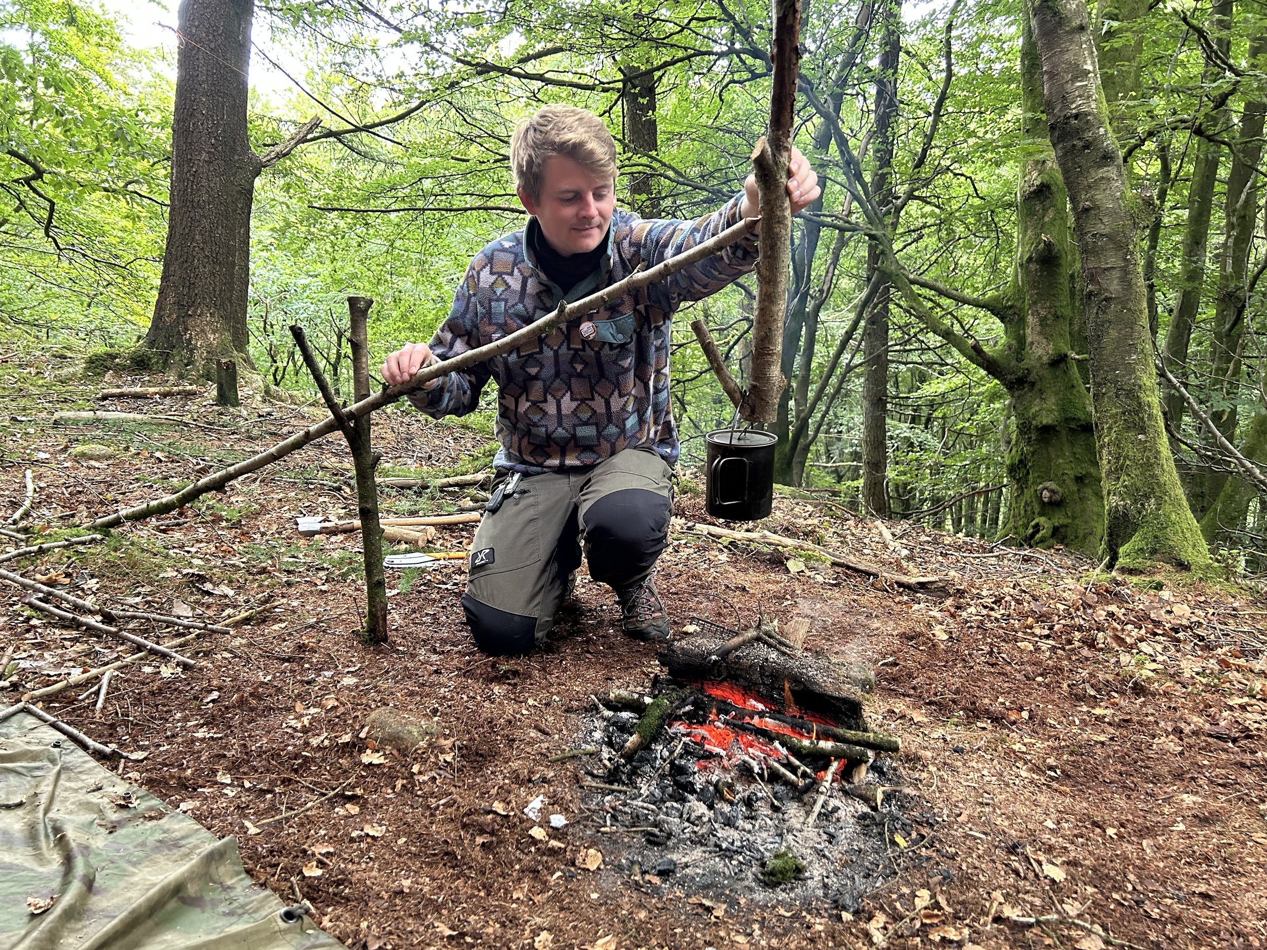 A person kneeling on the forest floor tending a small campfire with red glowing embers. The person is wearing outdoor gear and is surrounded by green trees and foliage. There are sticks and a black mug nearby.