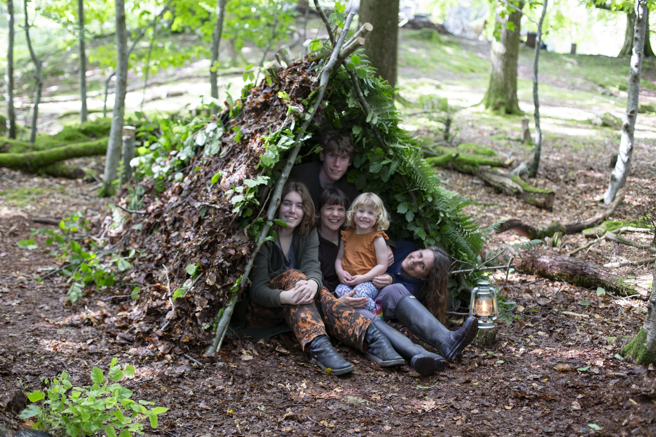 Group of five people, three adults and two children, sitting inside a makeshift shelter made of leaves and sticks in a forest. There is a lantern on the ground nearby.