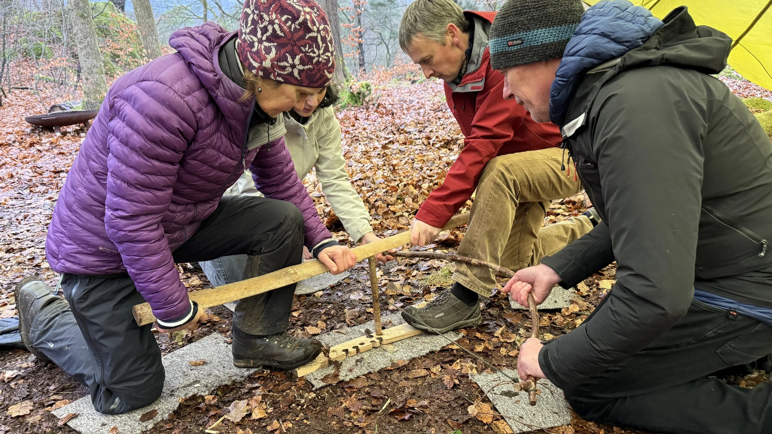 Bushcraft in the Lake District. Four people wearing outdoor clothing are crouched on the ground in a wooded area, working together to make an ember using group bow drill friction fire. The ground is covered with fallen leaves.