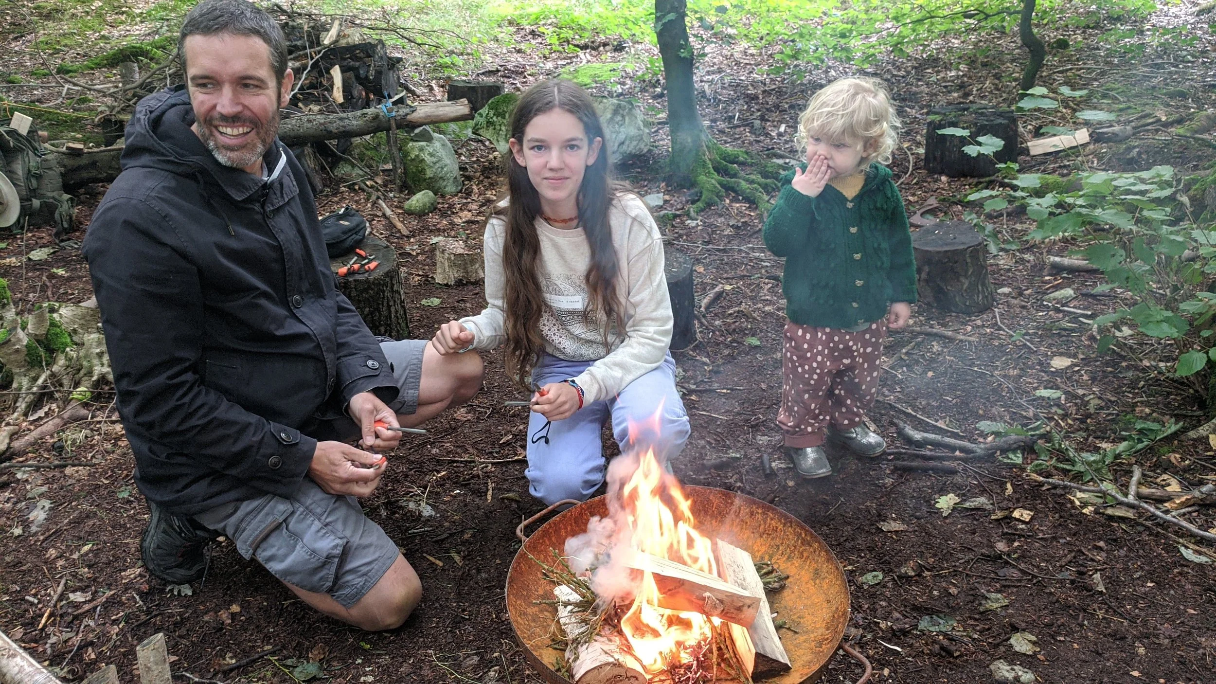 Bushcraft in the Lake District. Man and two young girls sitting and standing around a small campfire in a wooded area, with trees and fallen logs in the background.