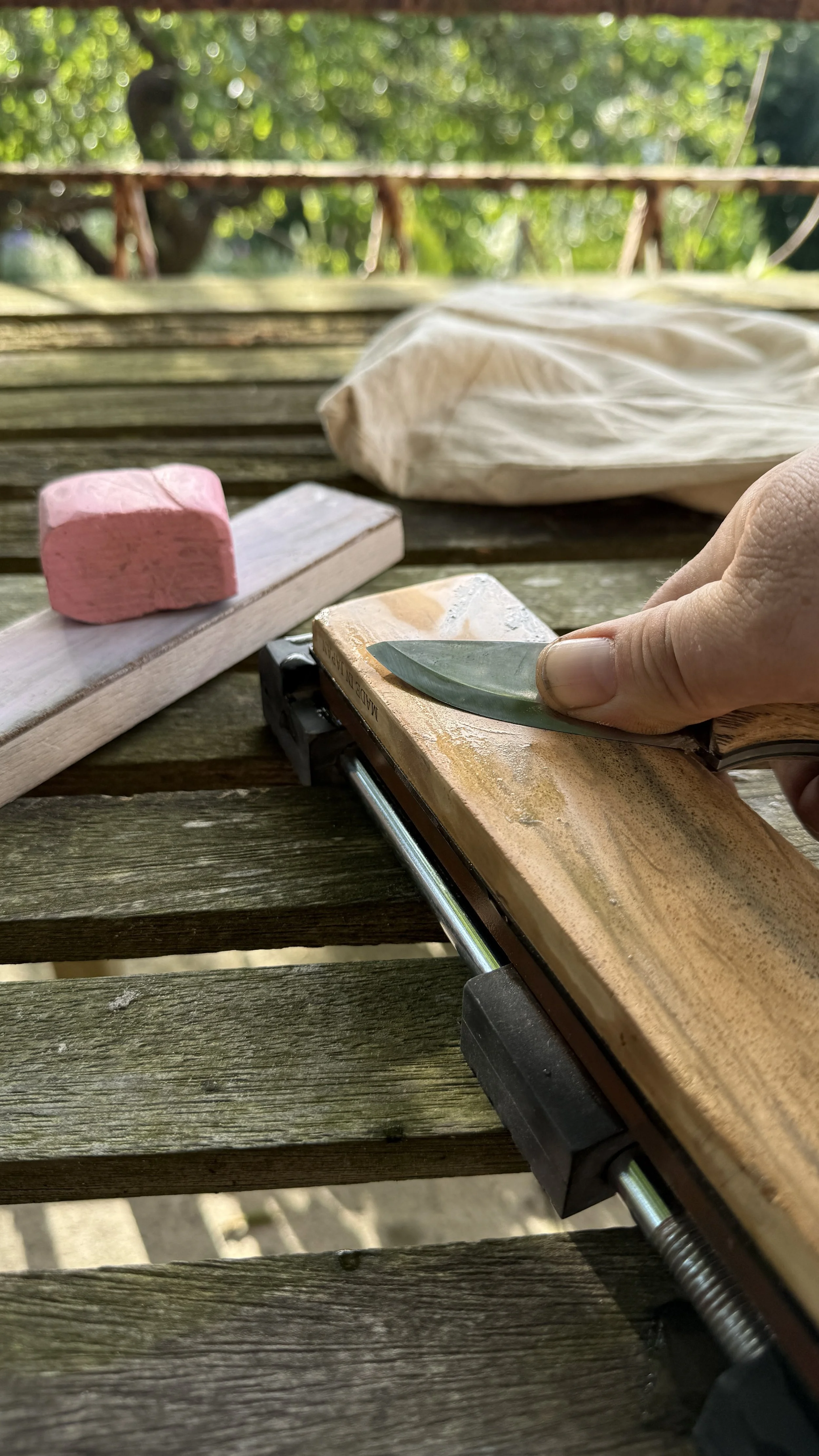 Bushcraft in the Lake District. A hand holding a small metal knife sharpening on a wetstone. Pink stropping compound and a leather strop are in the background, outdoors with green foliage.