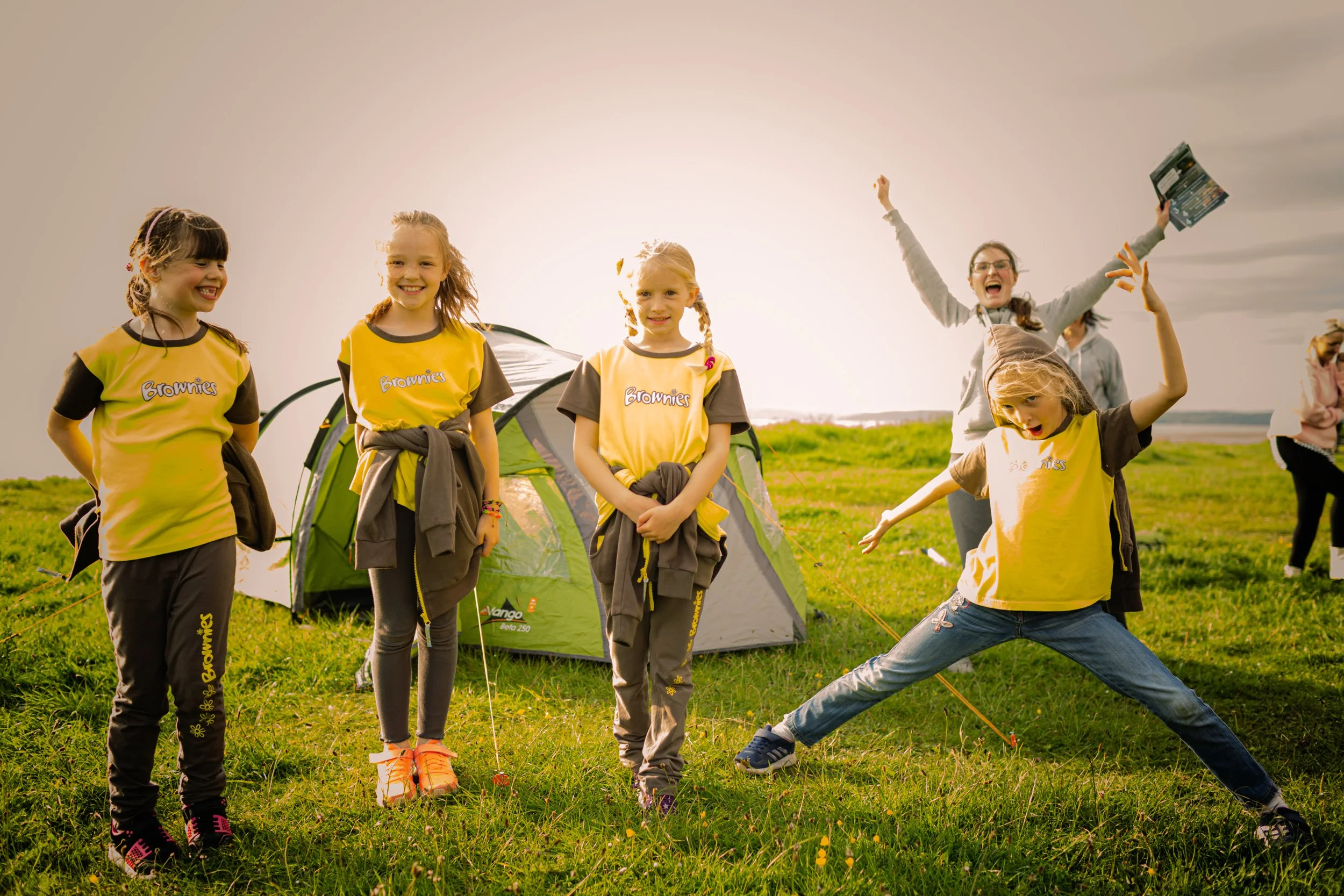 Bushcraft in the Lake District. Group of children and a woman camping outdoors on a grassy field with tents, smiling and playing together during daytime.