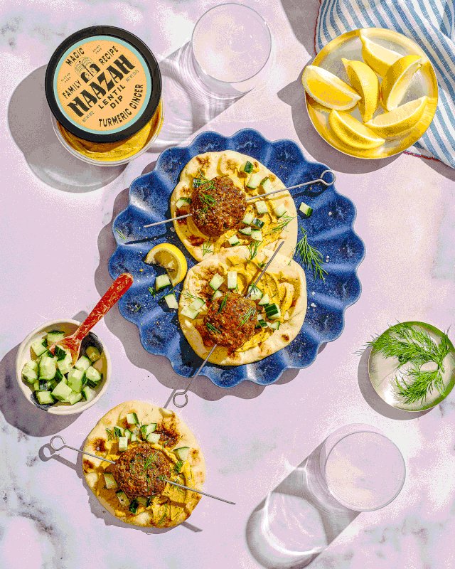 Hummus with meatballs, cucumbers, and herbs on a blue plate, surrounded by lemon wedges, a jar of turmeric garlic dip, a bowl of chopped cucumbers. Maazah Dips. 
