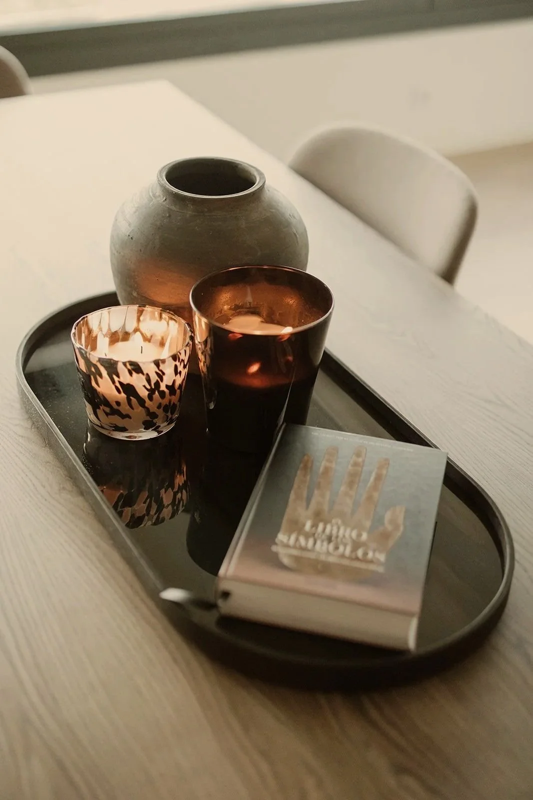 A black tray on a white table holds a grey ceramic vase, a lit candle in a leopard print holder, a dark drink in a glass, and a matchbook with a raised hand symbol on the cover.